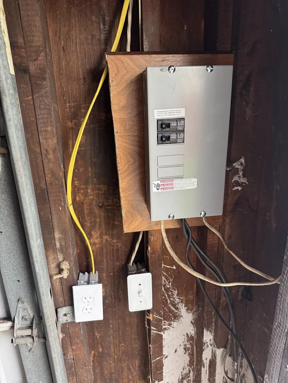 Electrical panel mounted on a wooden wall with yellow and black power cords connected, and a white wall outlet nearby.