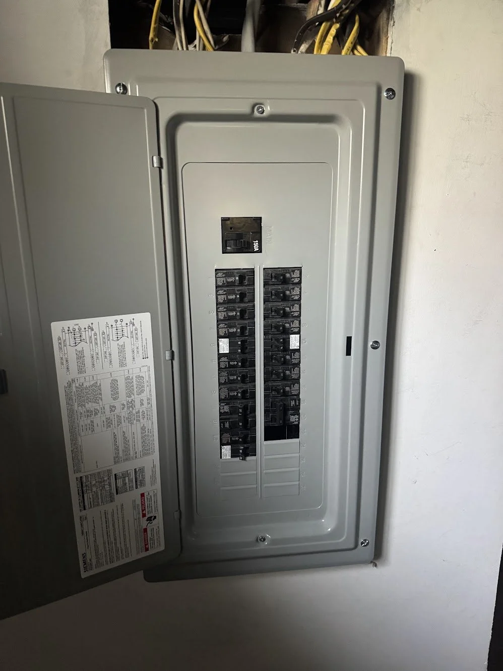 An electrical circuit breaker panel mounted on a wall, with the door open revealing various circuit breakers inside and electrical wiring above.
