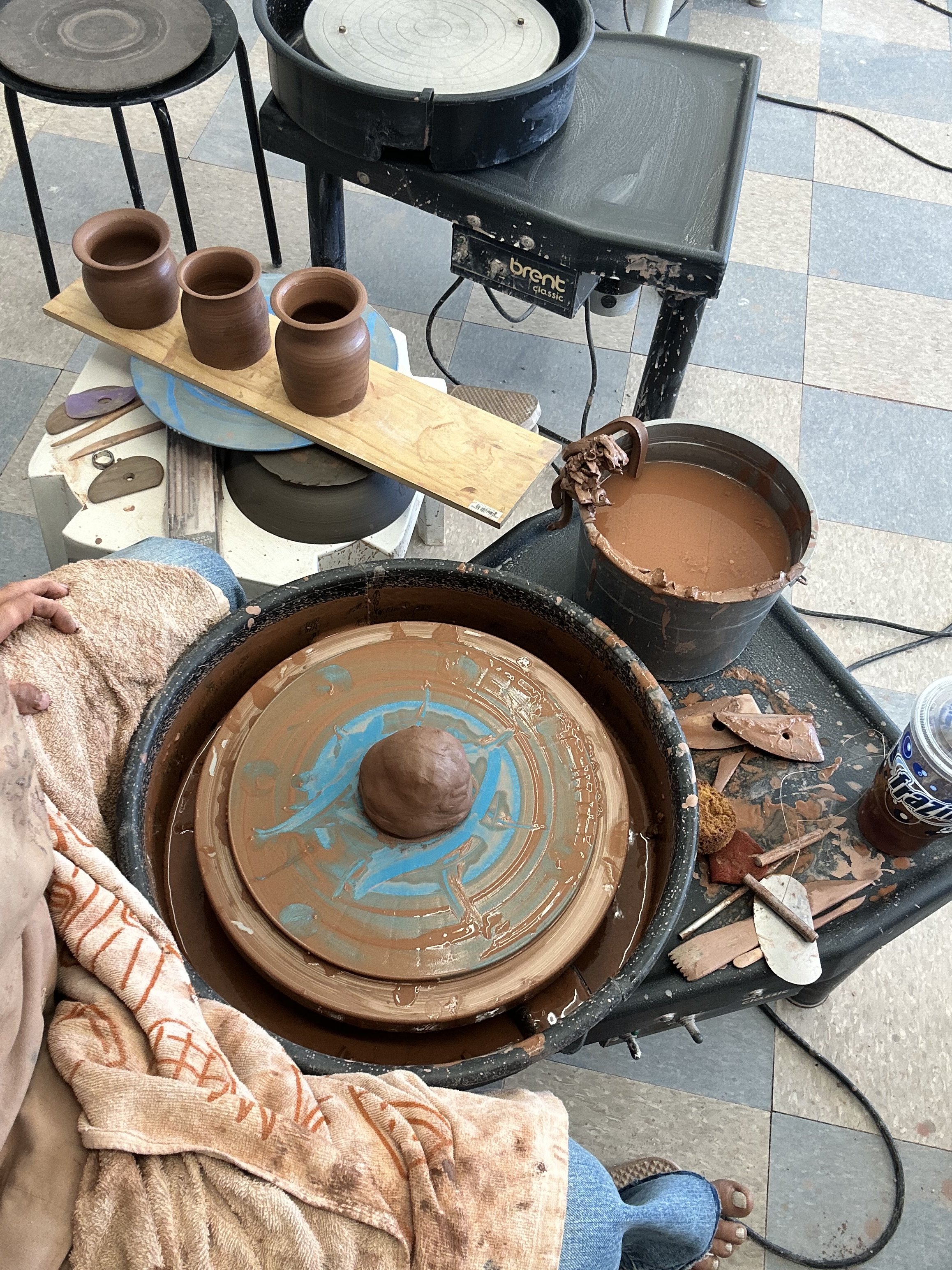 A workspace with a pottery wheel in use, small clay pots, buckets of clay, and various pottery tools on a tiled floor.