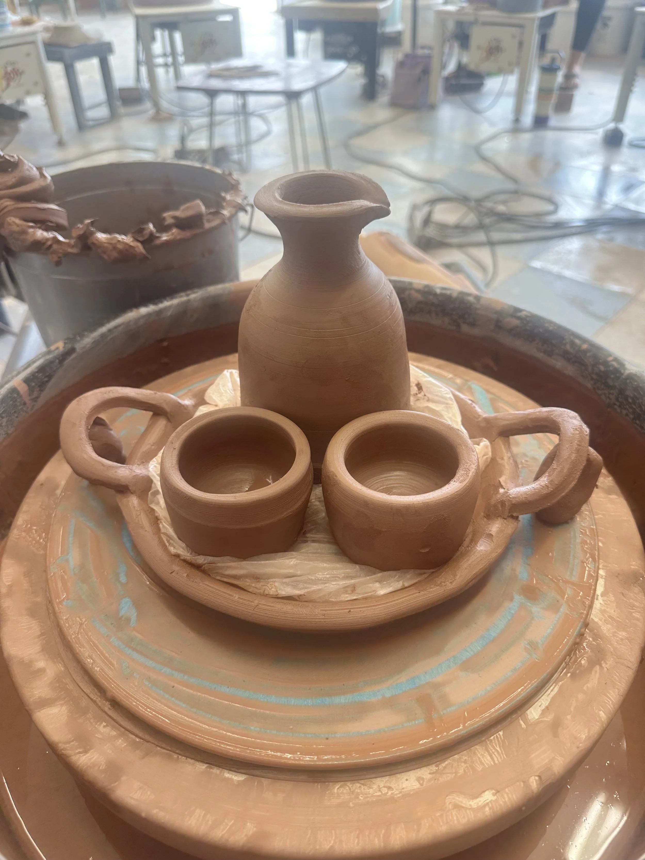 Unfired clay pottery on a pottery wheel, including a small vase and several cups, in a pottery studio with tables and tools in the background.