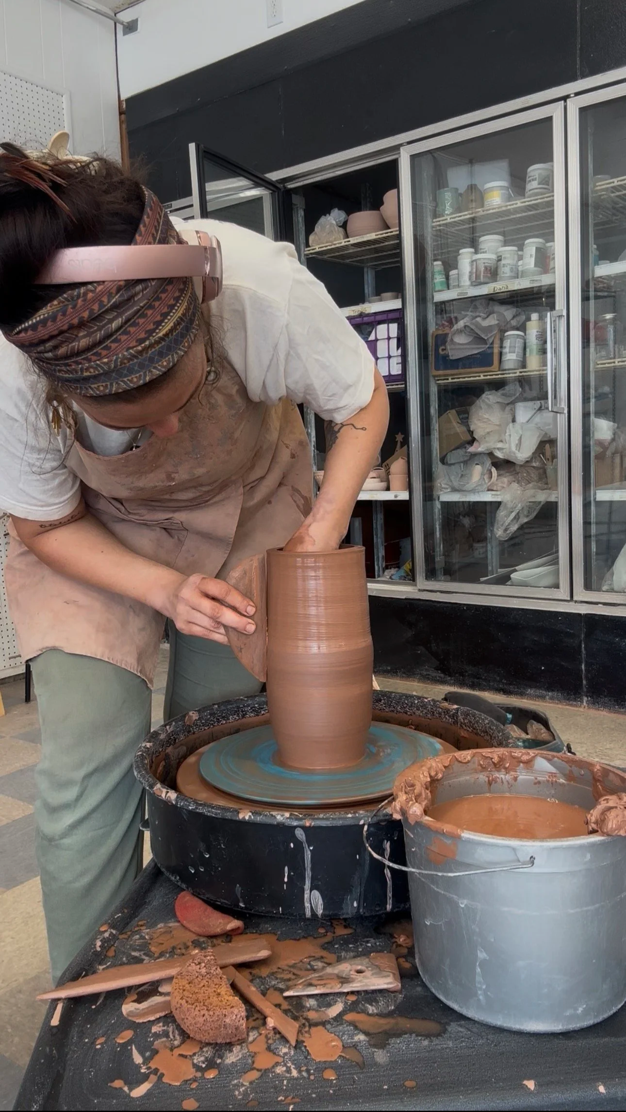 A person shaping a tall ceramic vase on a pottery wheel in a workshop, surrounded by pottery tools and buckets of clay.