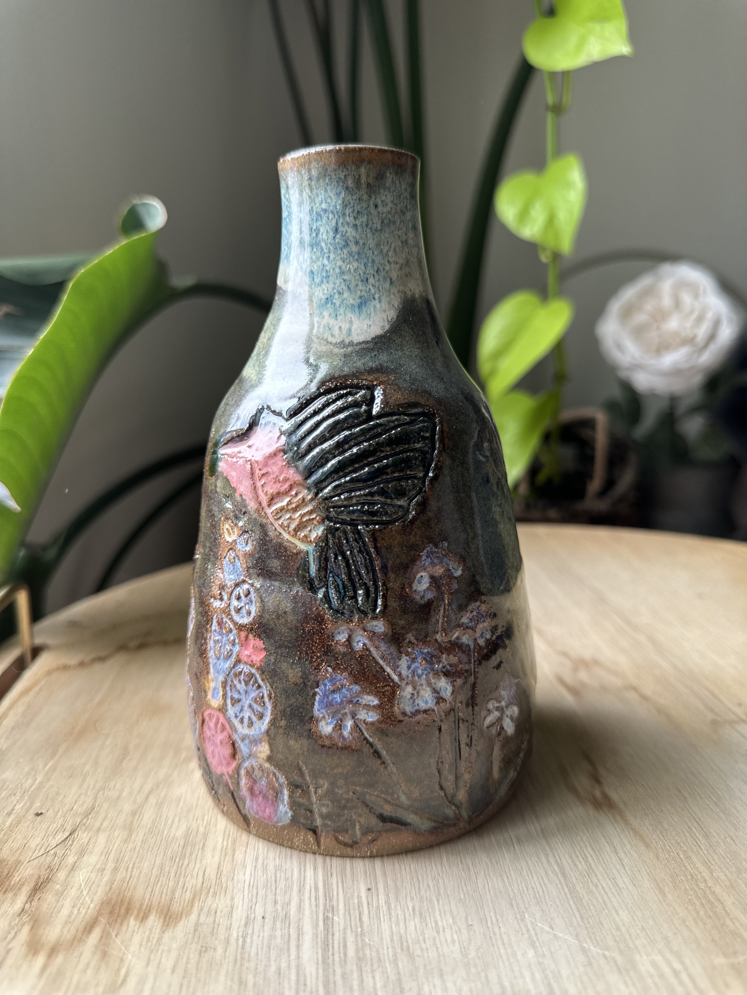 A ceramic vase with textured floral design and multicolored glaze, placed on a wooden surface with green houseplants in the background.