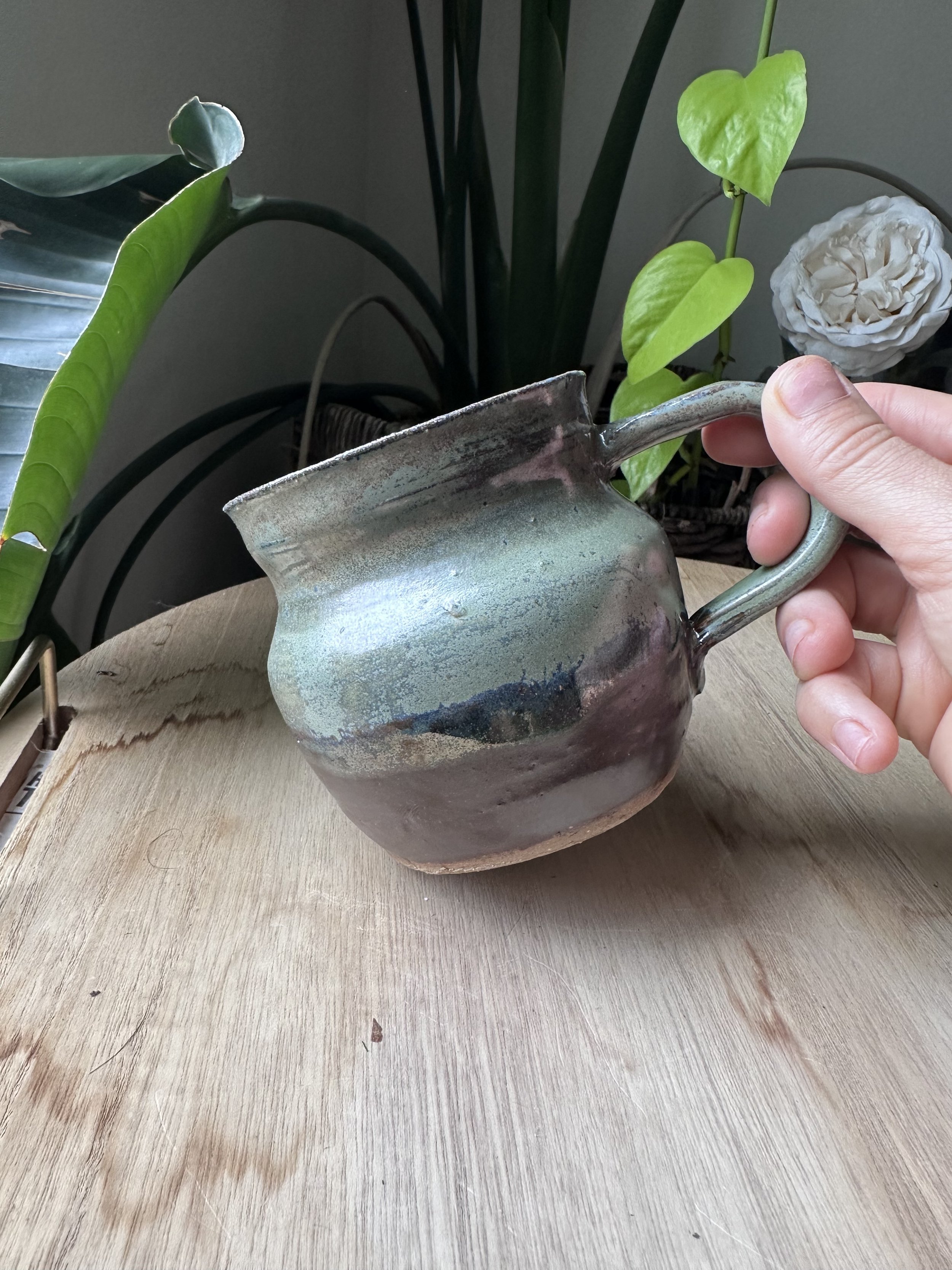 A person holding a ceramic watering can with a greenish glaze, placed on a wooden table, with green leafy plants and a white decorative flower in the background.
