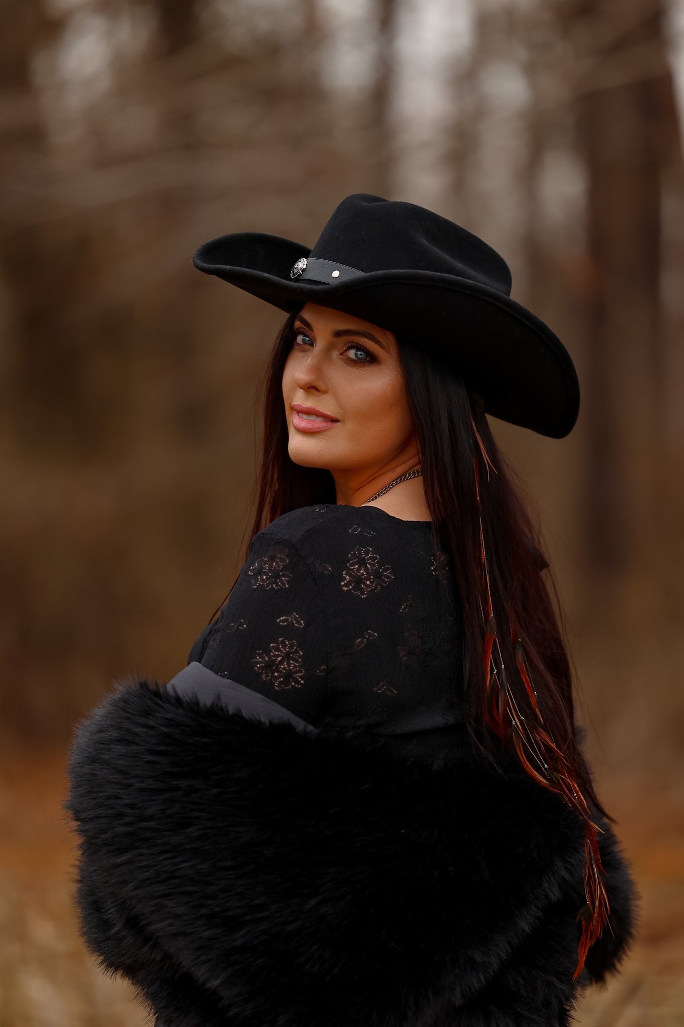 A woman with long dark hair and blue eyes wearing a large black cowboy hat with a decorative band, a black lace top, and a furry black coat, outdoors in a blurred natural setting with trees in the background.