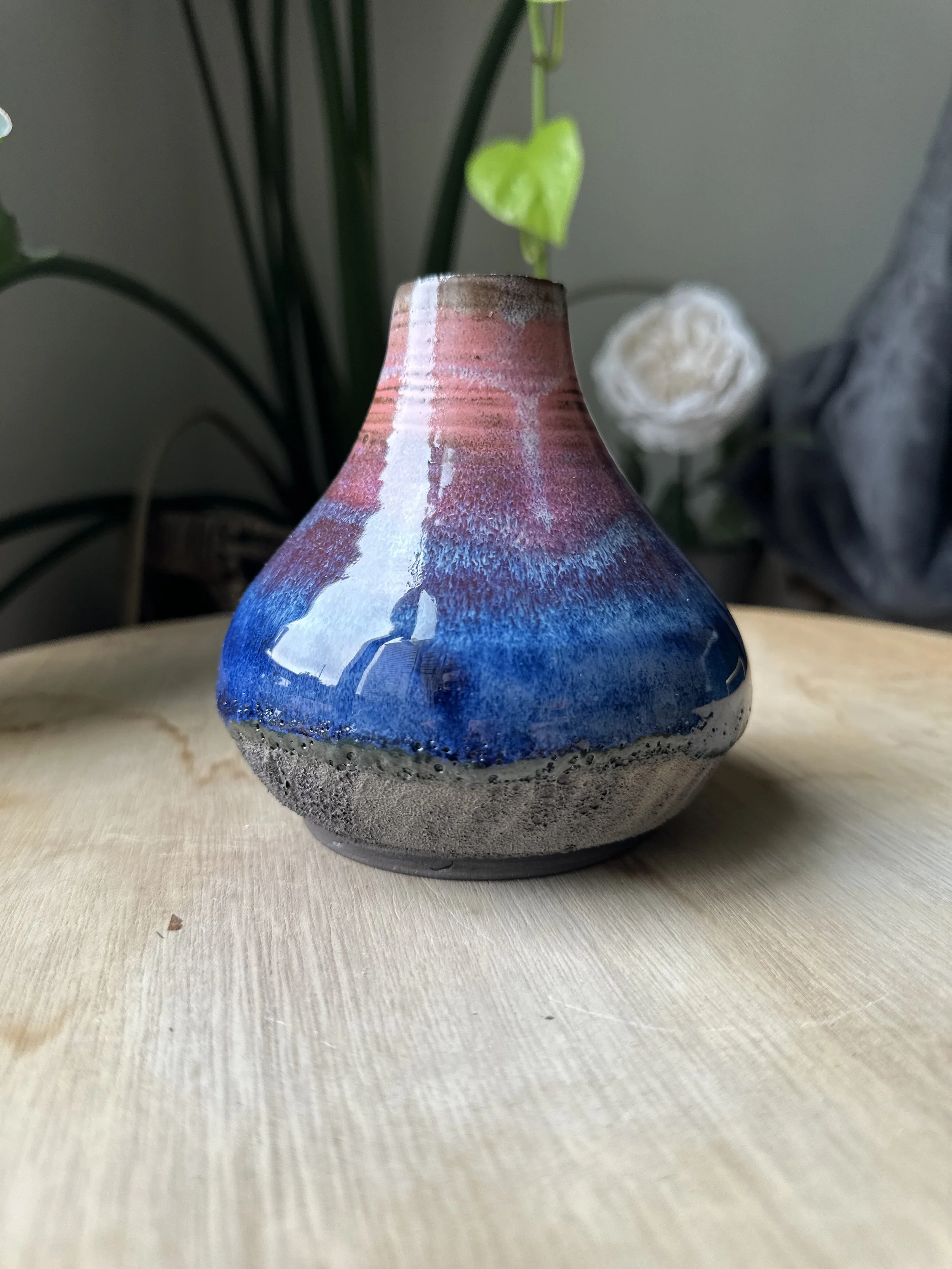 A ceramic vase with a colorful glaze featuring shades of pink, purple, and blue, sitting on a light-colored wooden surface with some green plants in the background.