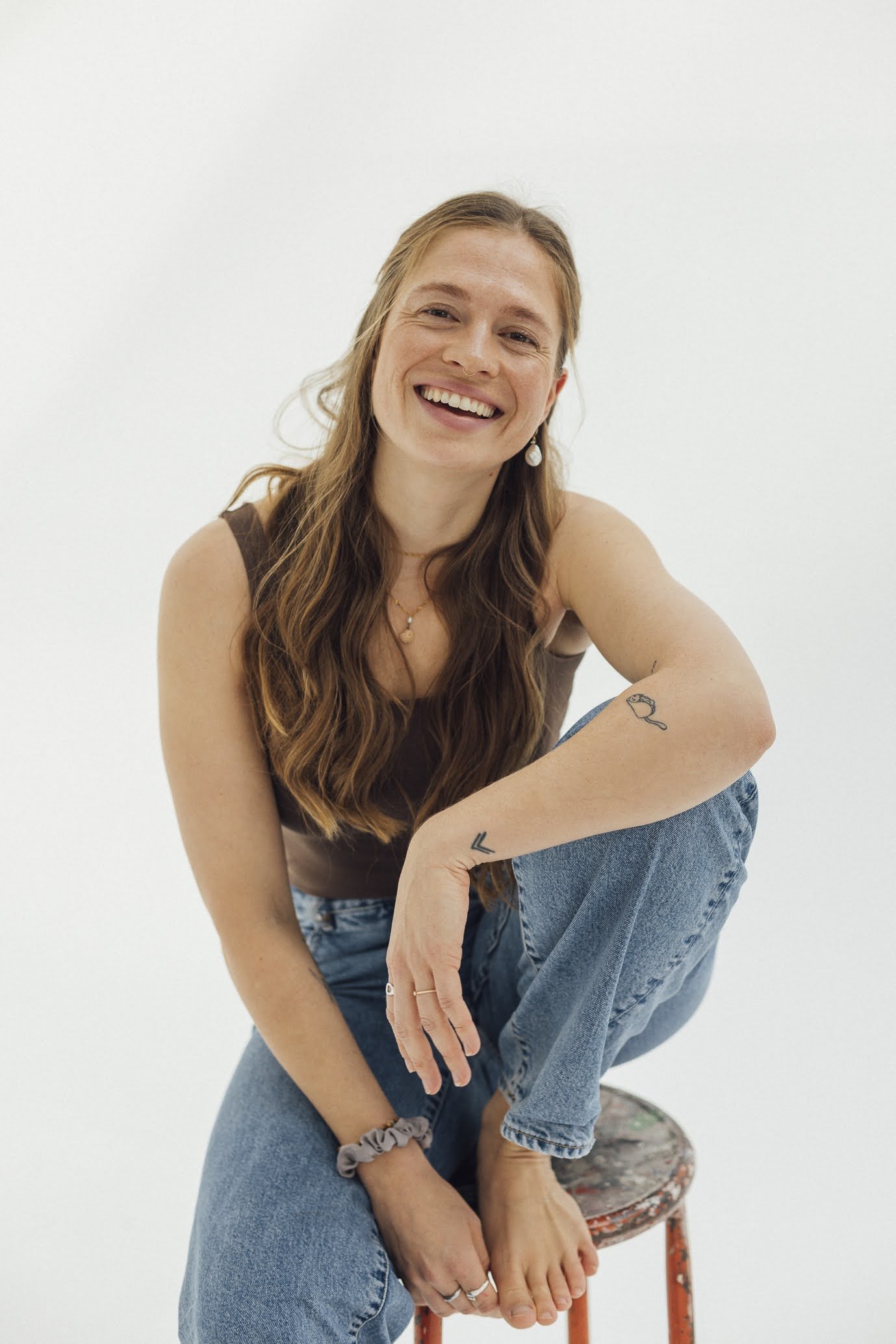 Junge Frau mit langen braunen Haaren, lachend, trägt ein braunes Top, blaue Jeans, sitzt auf einem gestreiften Hocker an weißem Hintergrund.