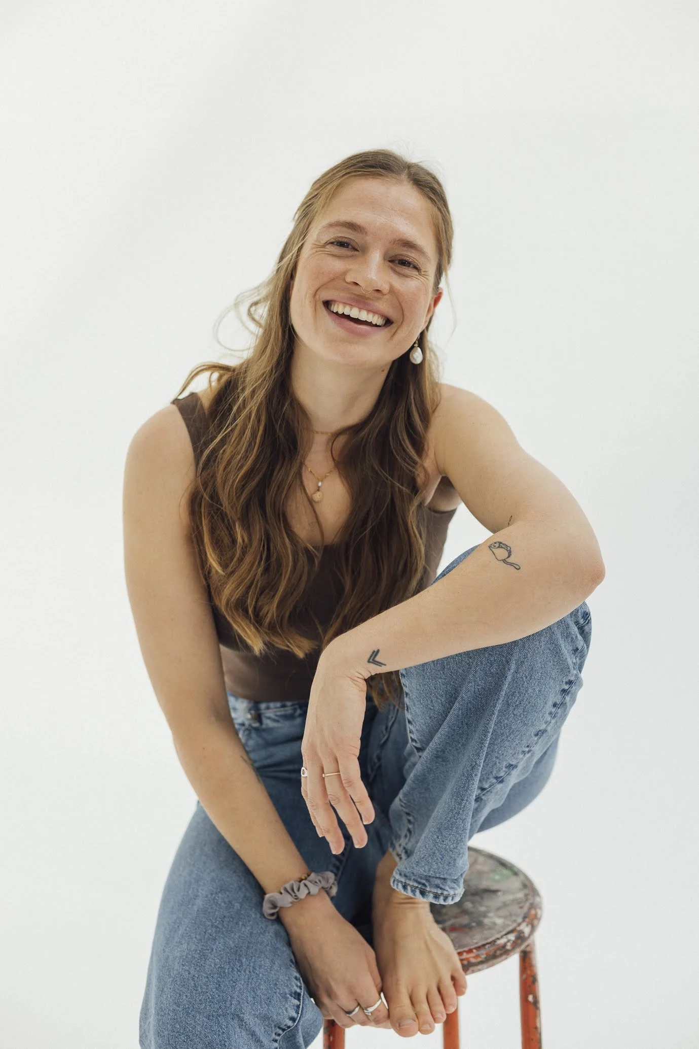 Junge Frau mit langen braunen Haaren, lachend, sitzt auf einem alten, roten Hocker, trägt einen braunen Top und blaue Jeans, tätowiert am Arm, trägt Schmuck, gegen weißen Hintergrund.
