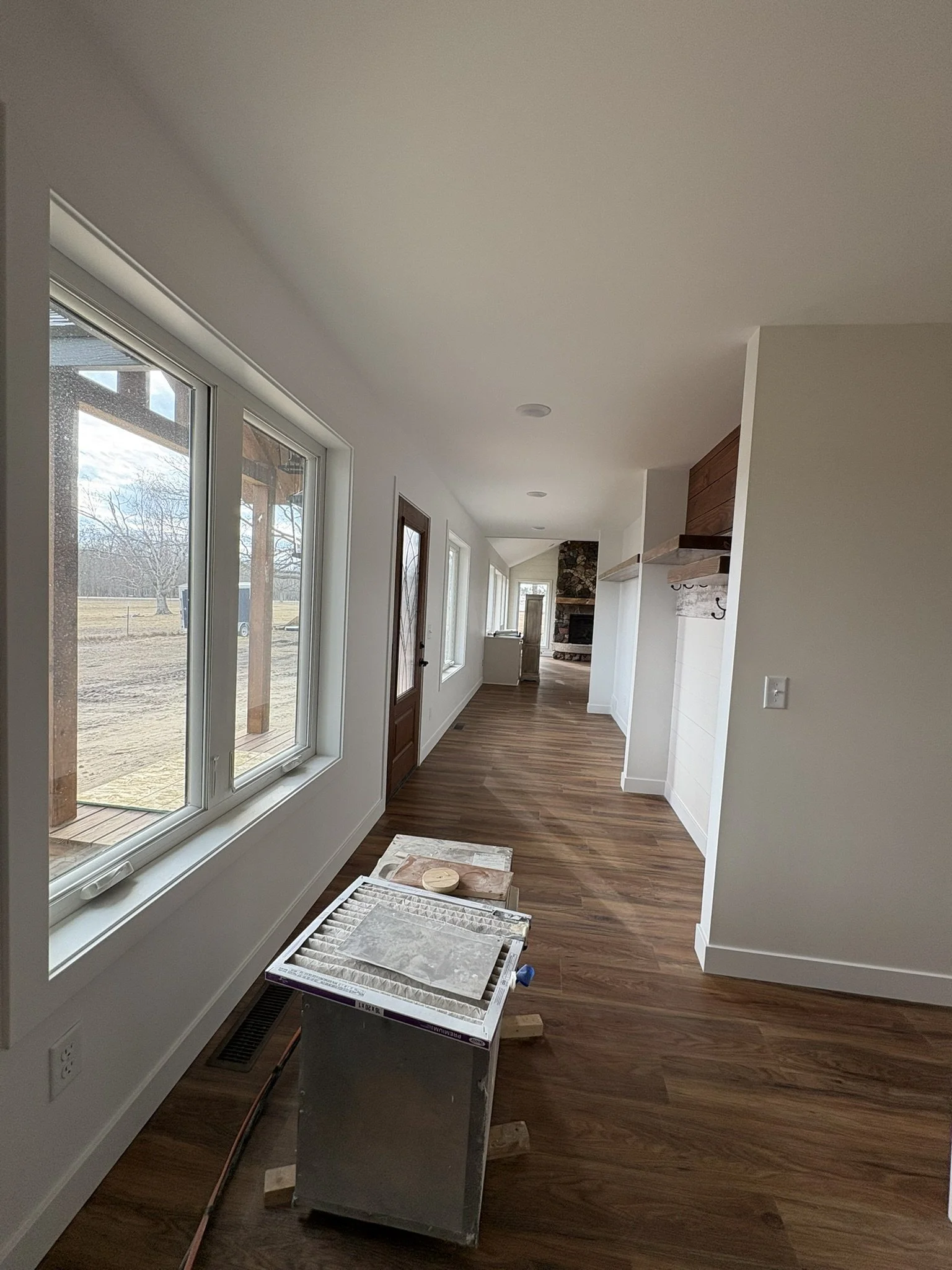 Interior view of a hallway with large windows, a stone fireplace at the end, and hardwood floors. There is a heating vent, an air filter, and some tools on the floor.