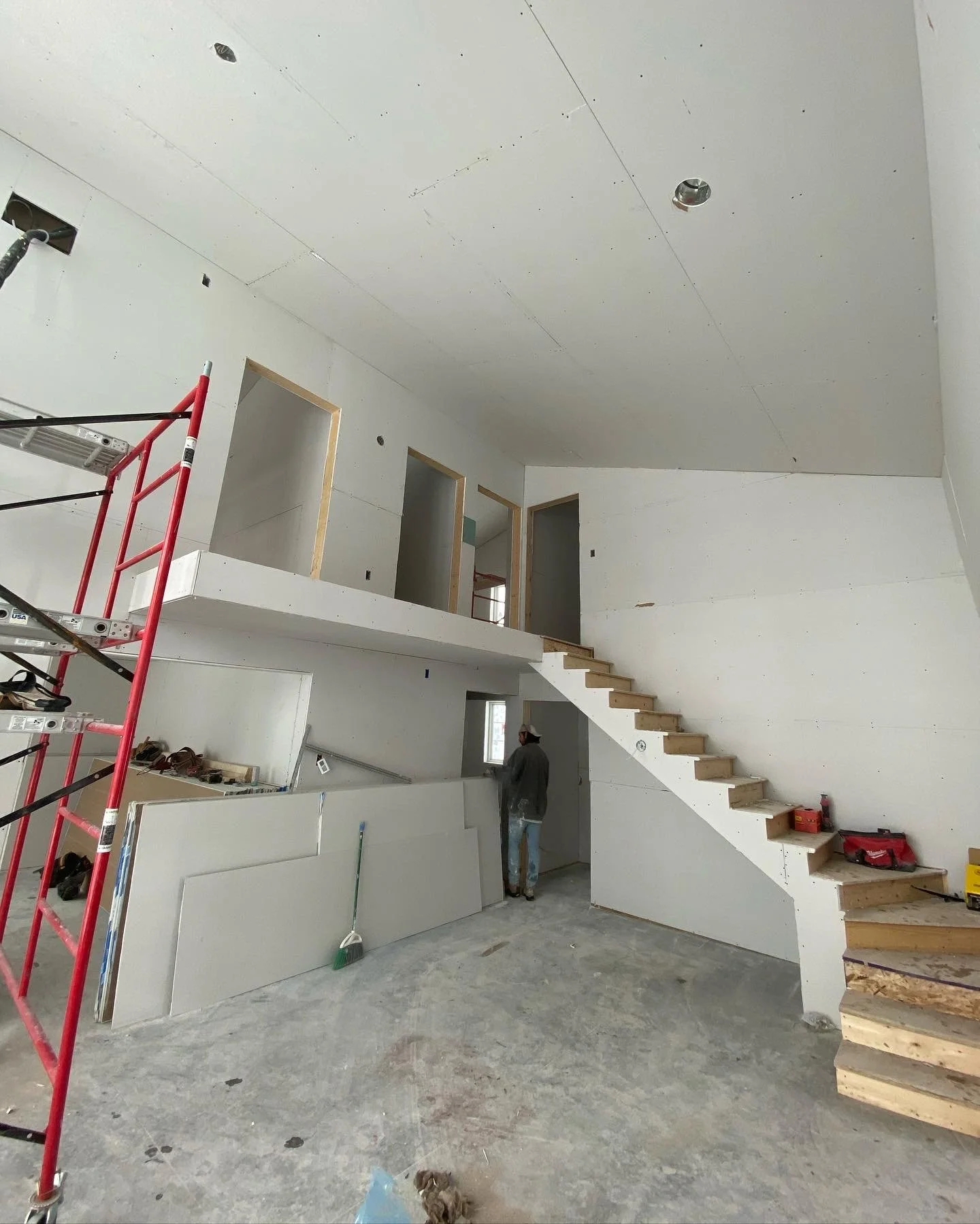Interior of a house under construction with drywall installation, scaffolding, staircase, and a worker.