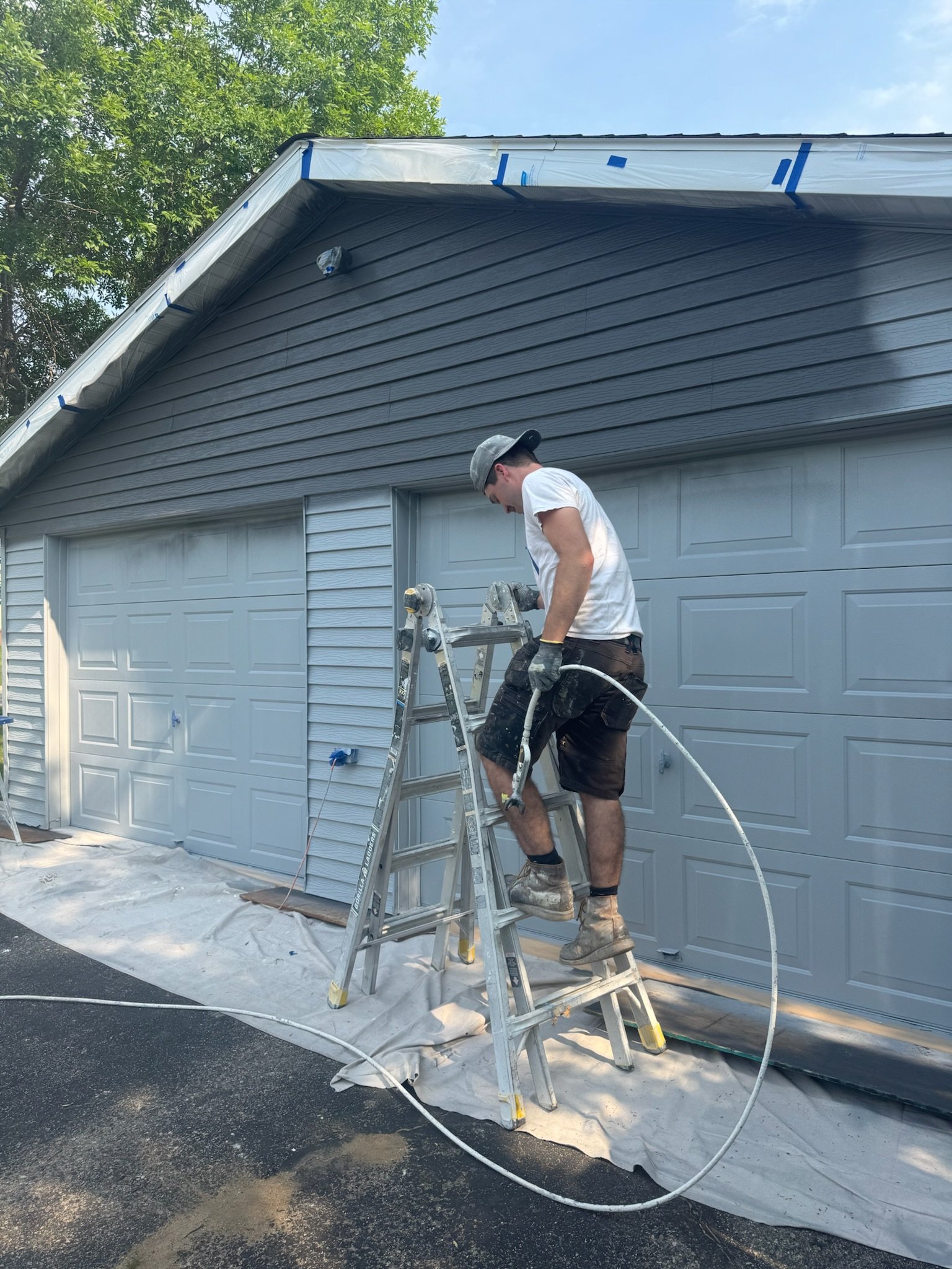 A man in a white t-shirt, brown shorts, and work boots is standing on a step ladder, painting the exterior of a house. The house has a double garage door and blue siding. He is wearing a gray cap and is holding a paint sprayer, with spray paint residue visible on the ground and a plastic drop cloth underneath.