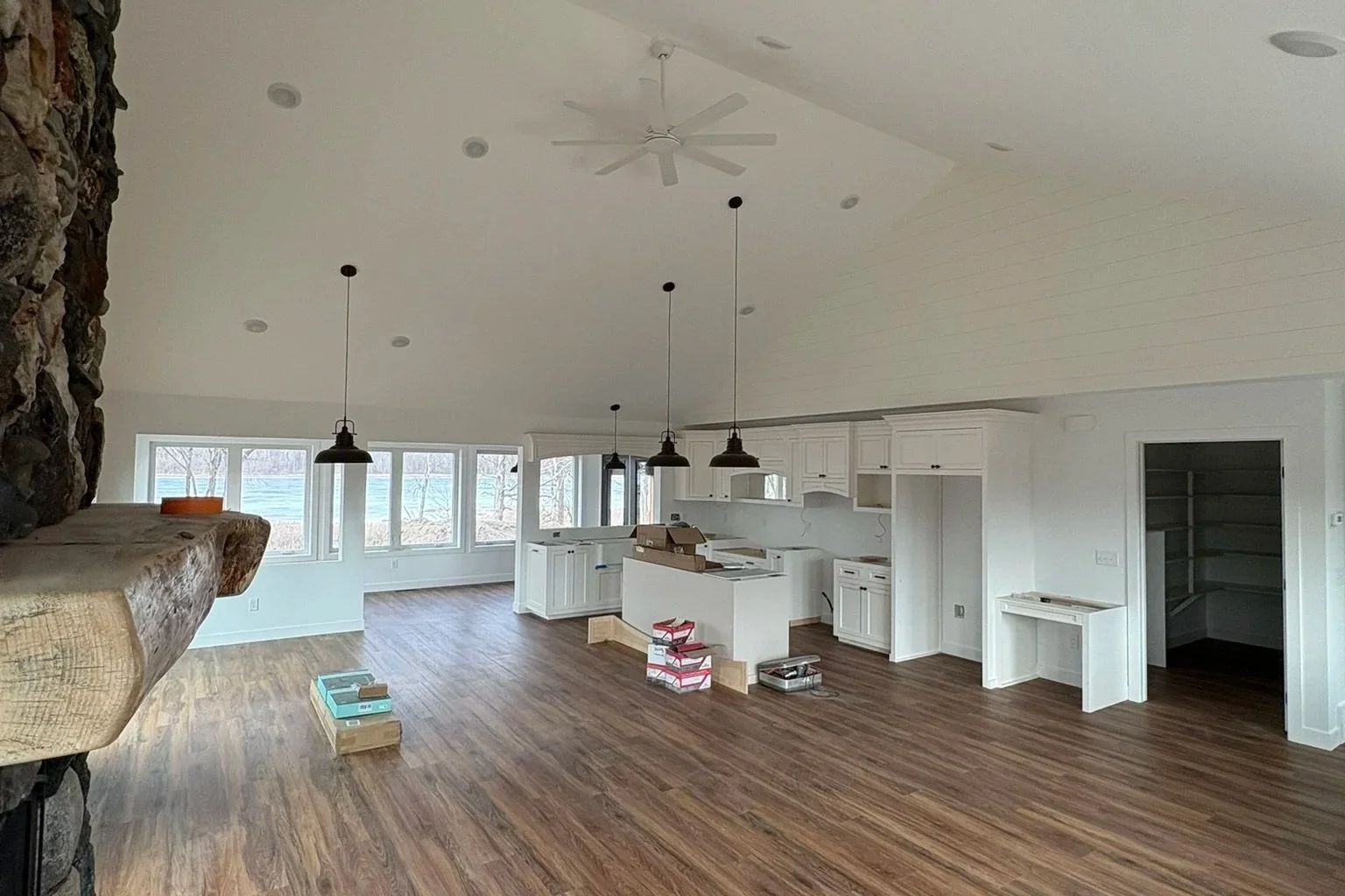 Empty living room with white kitchen cabinets, wooden floors, and large windows showing an outdoor view, under a white ceiling with a fan and pendant lights.