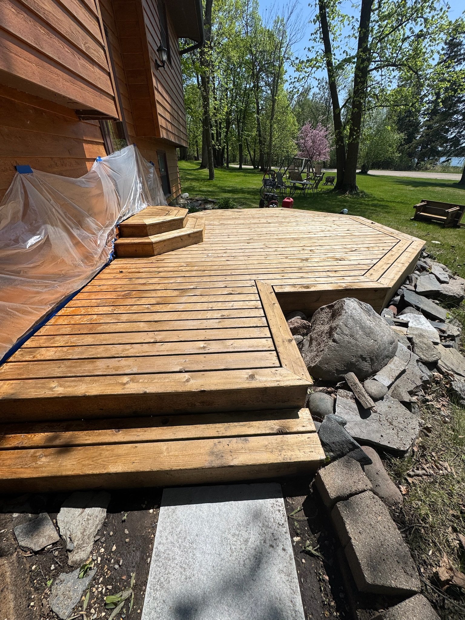 A wooden deck under construction near a house with brown siding. The deck has stairs and rocks around its edge. The yard is grassy with trees, and outdoor furniture and children's toys are visible in the background.