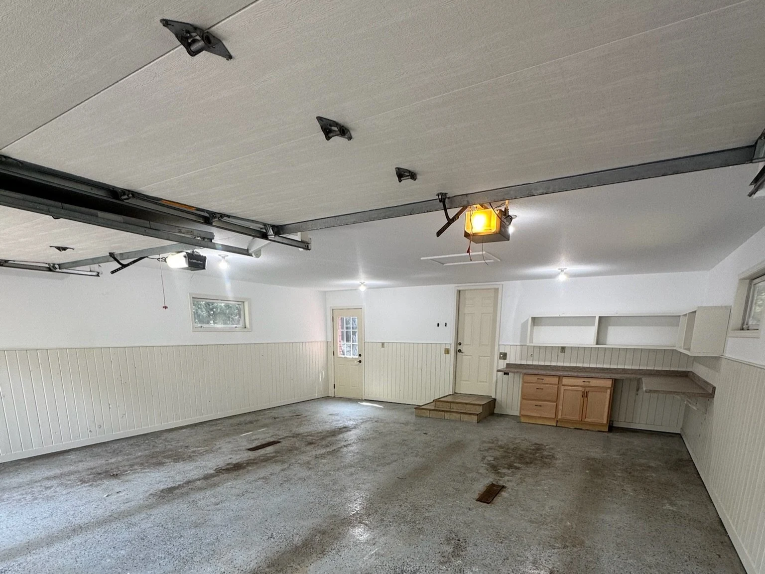 Empty garage with unfinished flooring, white walls, overhead garage door mechanism, small window, and built-in shelving