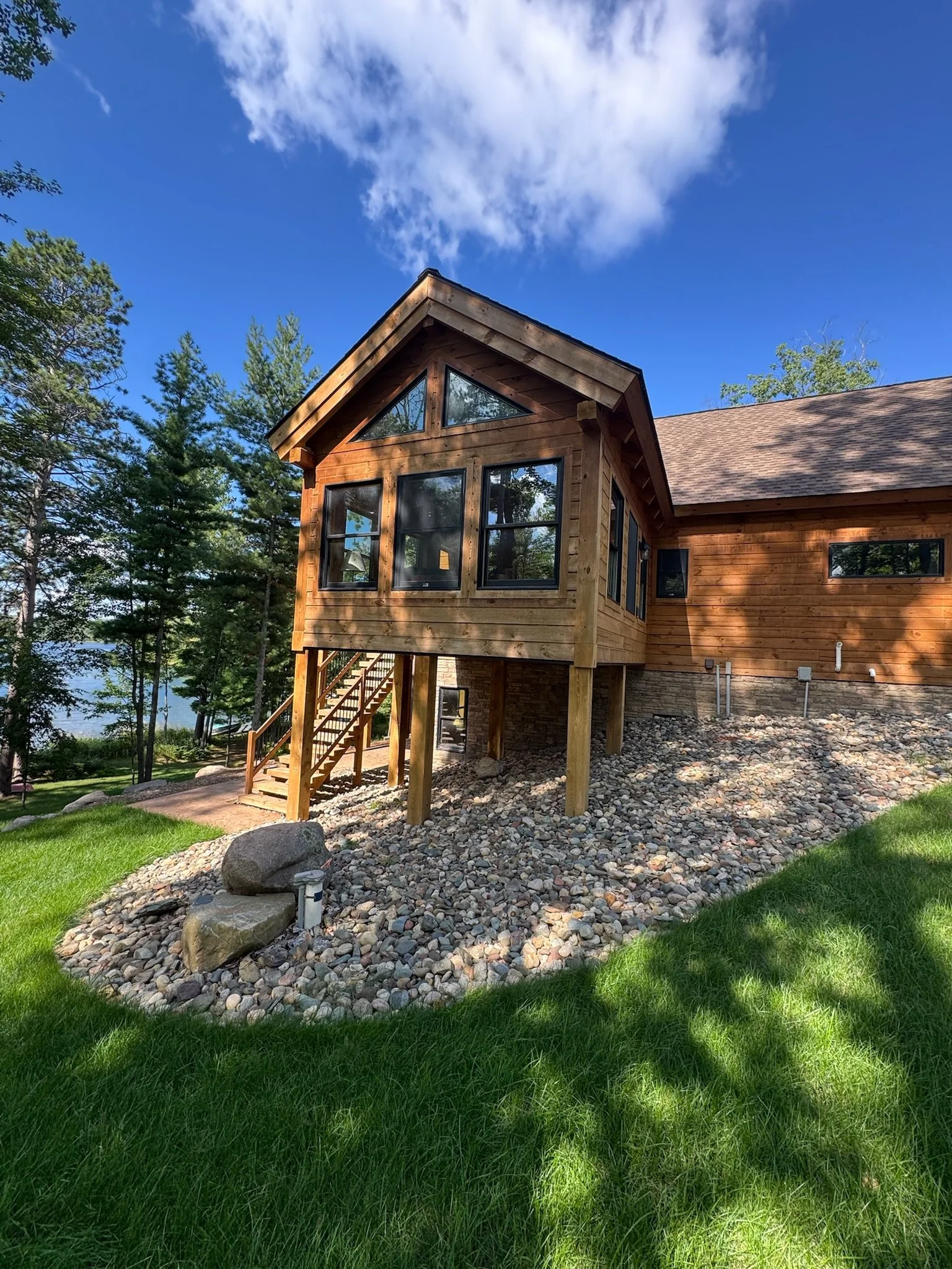 A wooden house with a steep gabled roof, multiple large windows, and a staircase leading up to the entrance, surrounded by green grass, trees, and a rocky landscape beneath a blue sky with clouds.