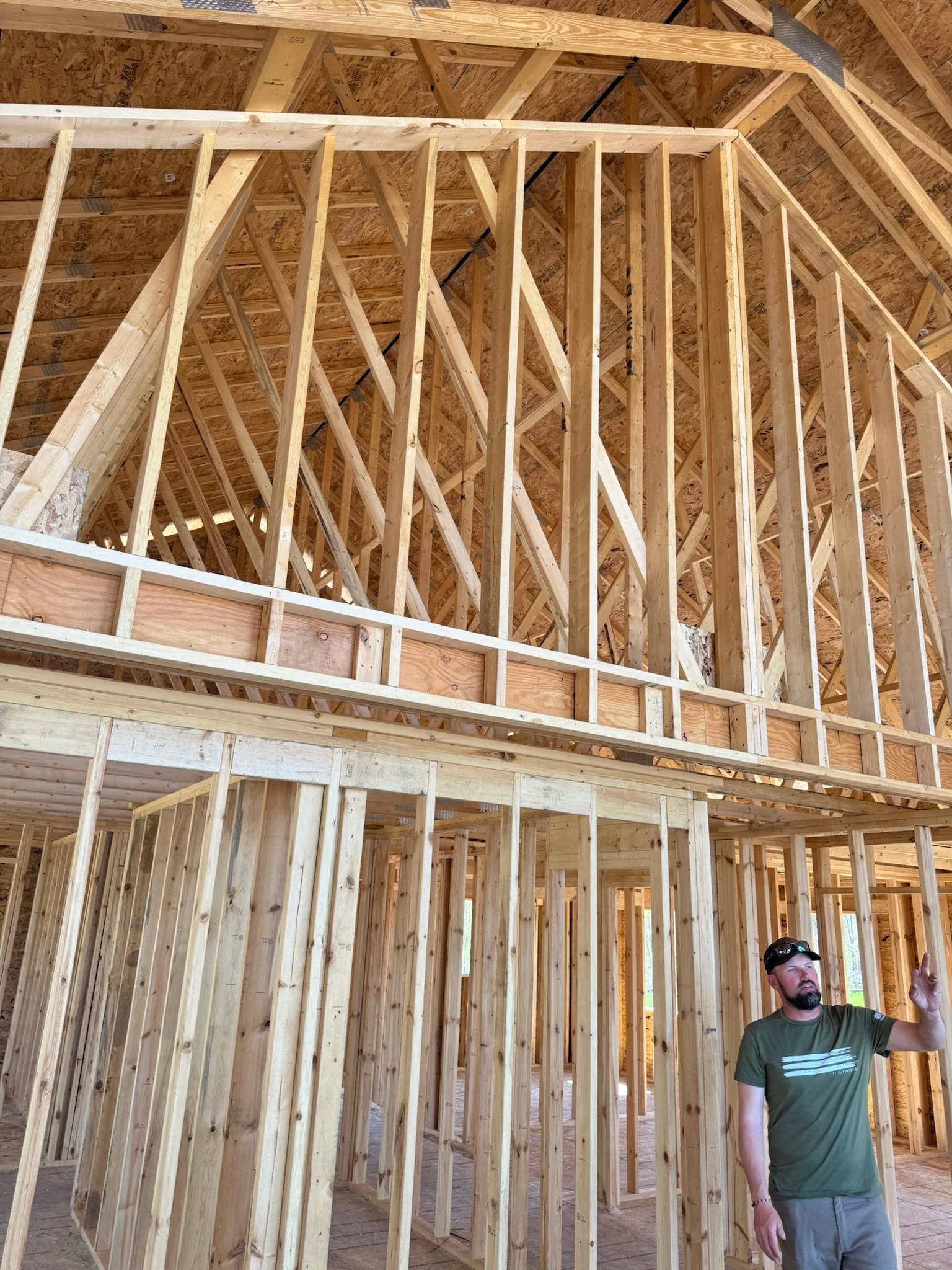 Interior view of a house under construction with exposed wooden framing and a man in a green shirt and cap speaking or explaining something.