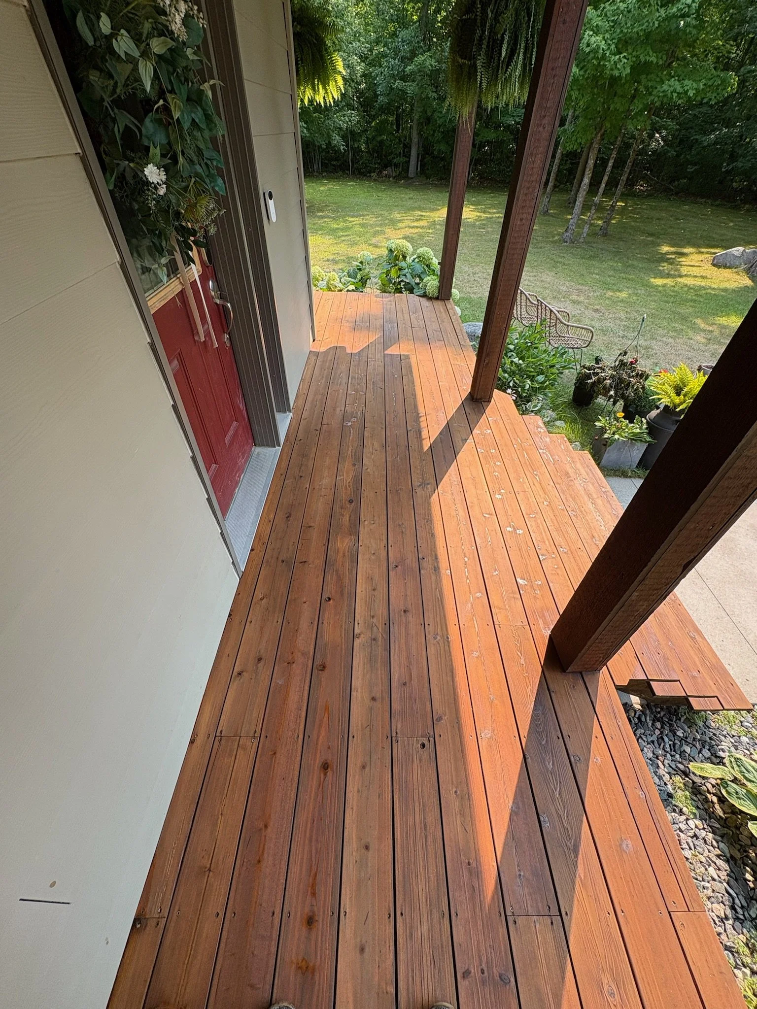 Wooden porch with stairs, shrubs, potted plants, and a backyard with grass and trees in the background.