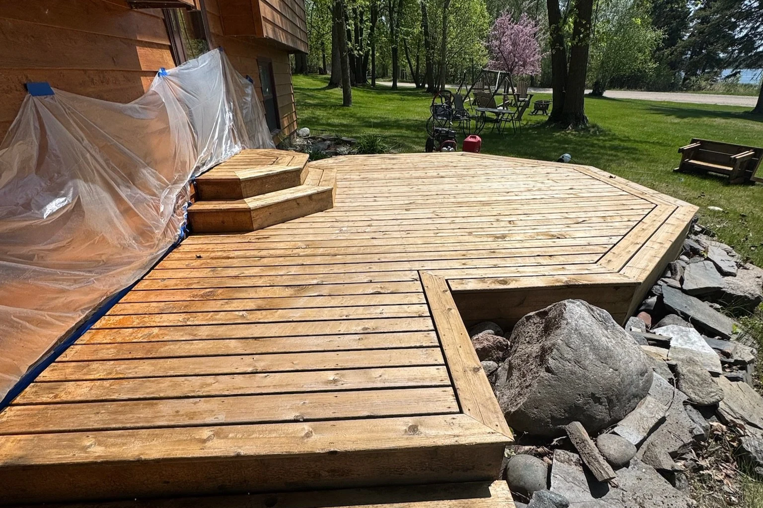 Wooden deck under construction with rocks surrounding it, outside of a house with lawn, trees, and outdoor furniture in the background.