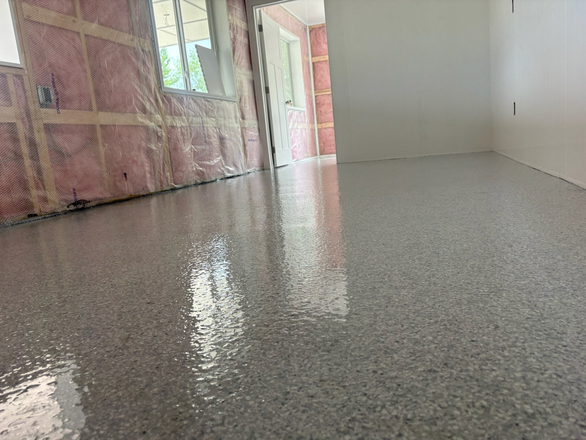 Interior of a house under construction, showing a polished concrete floor, unfinished walls with pink insulation and wooden framing, a white door, and windows providing natural light.