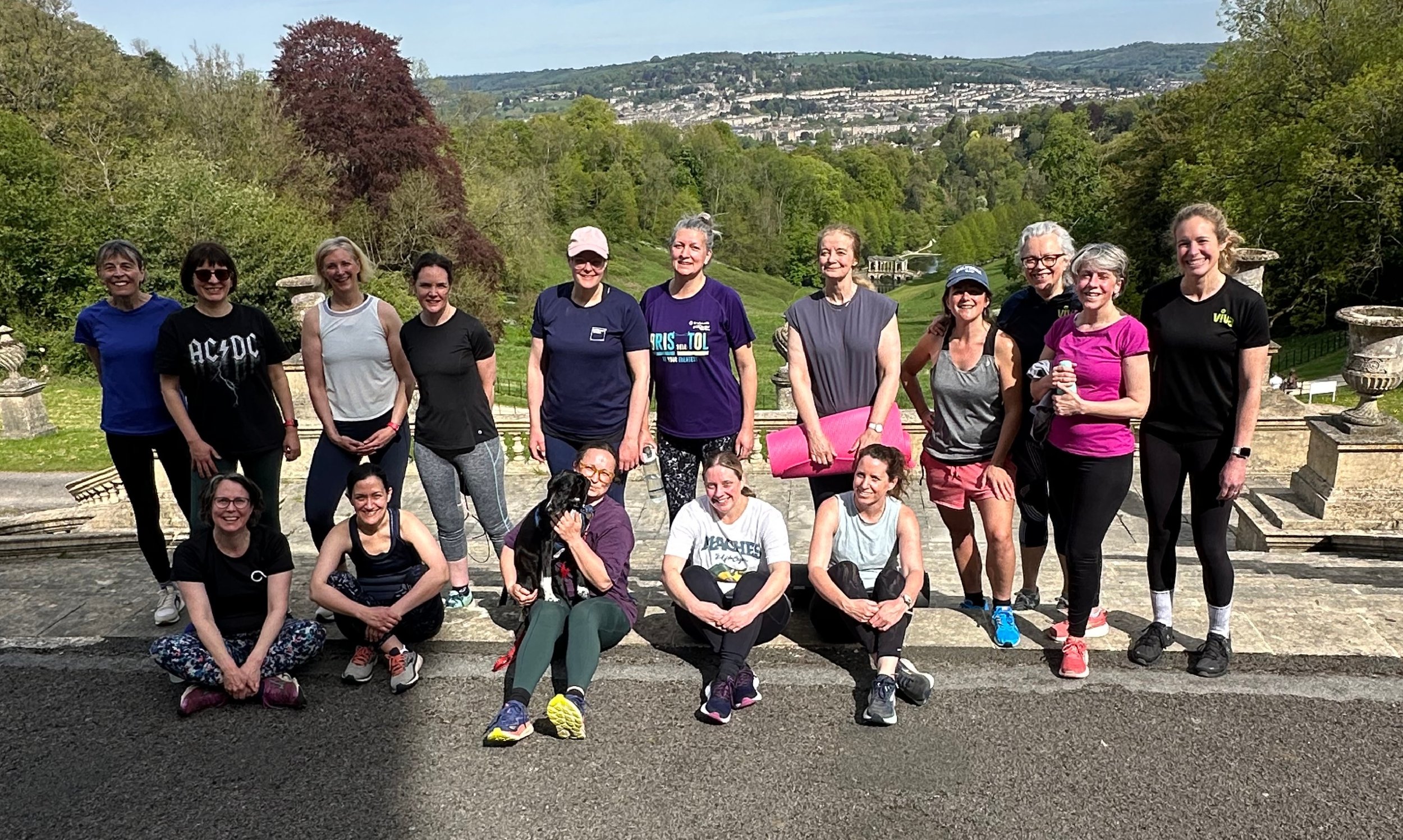 Group of women in athletic clothing posing outdoors on a stone staircase with a lush green hill and Bath cityscape in the background.