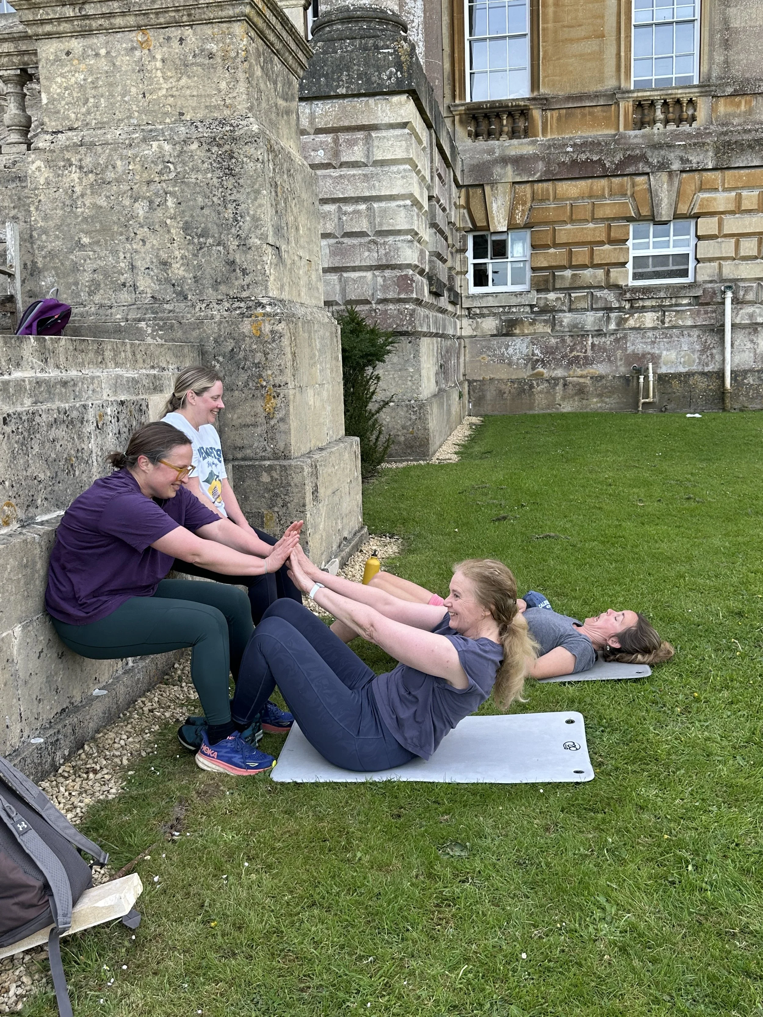 Two women are doing sit-up exercises outside on a grassy area, with one woman holding the ankles of the other woman as she exercises. They are next to a large stone building with windows, and a purple backpack is on the stone steps nearby.