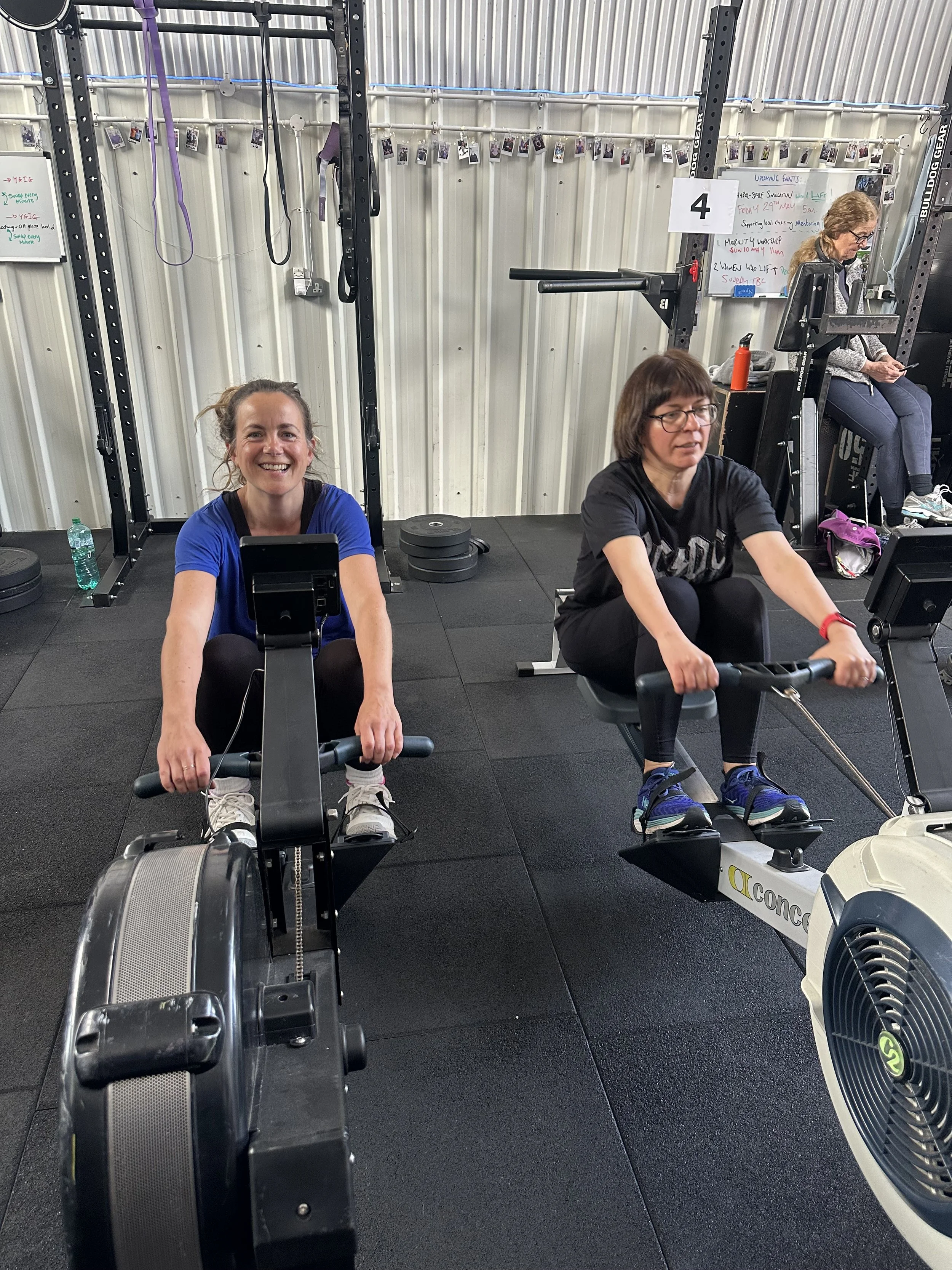Two women work out on rowing machines in a gym, with a third woman sitting on a weightlifting machine in the background. The gym has gray metal walls, a string of photos hanging above, and whiteboards with workouts written on them.