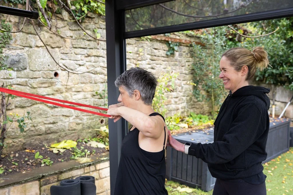 A woman performing resistance band exercises with assistance from another woman outdoors in a garden area.
