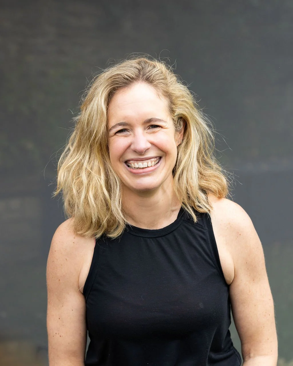 A woman with blonde, wavy hair smiling, wearing a sleeveless black top, standing outdoors with a blurred green and gray background.
