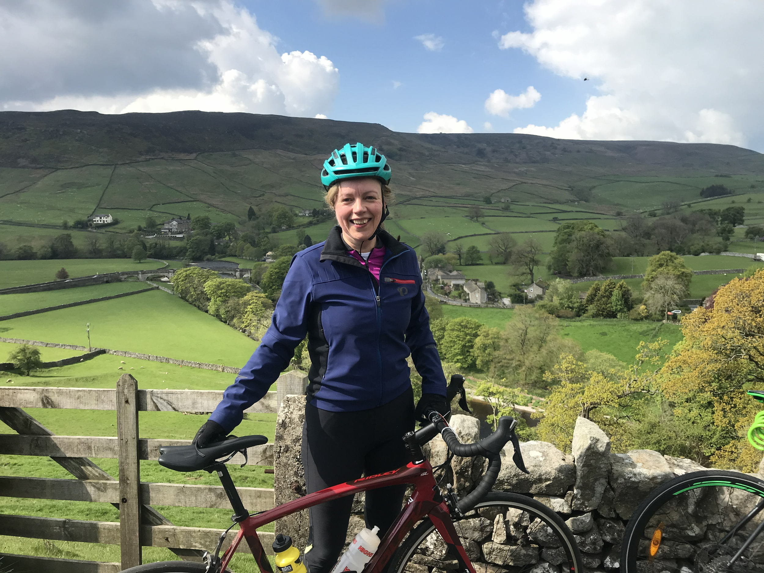 A smiling woman with a bicycle standing outdoors with green hills, trees, and a blue sky with clouds in the background.