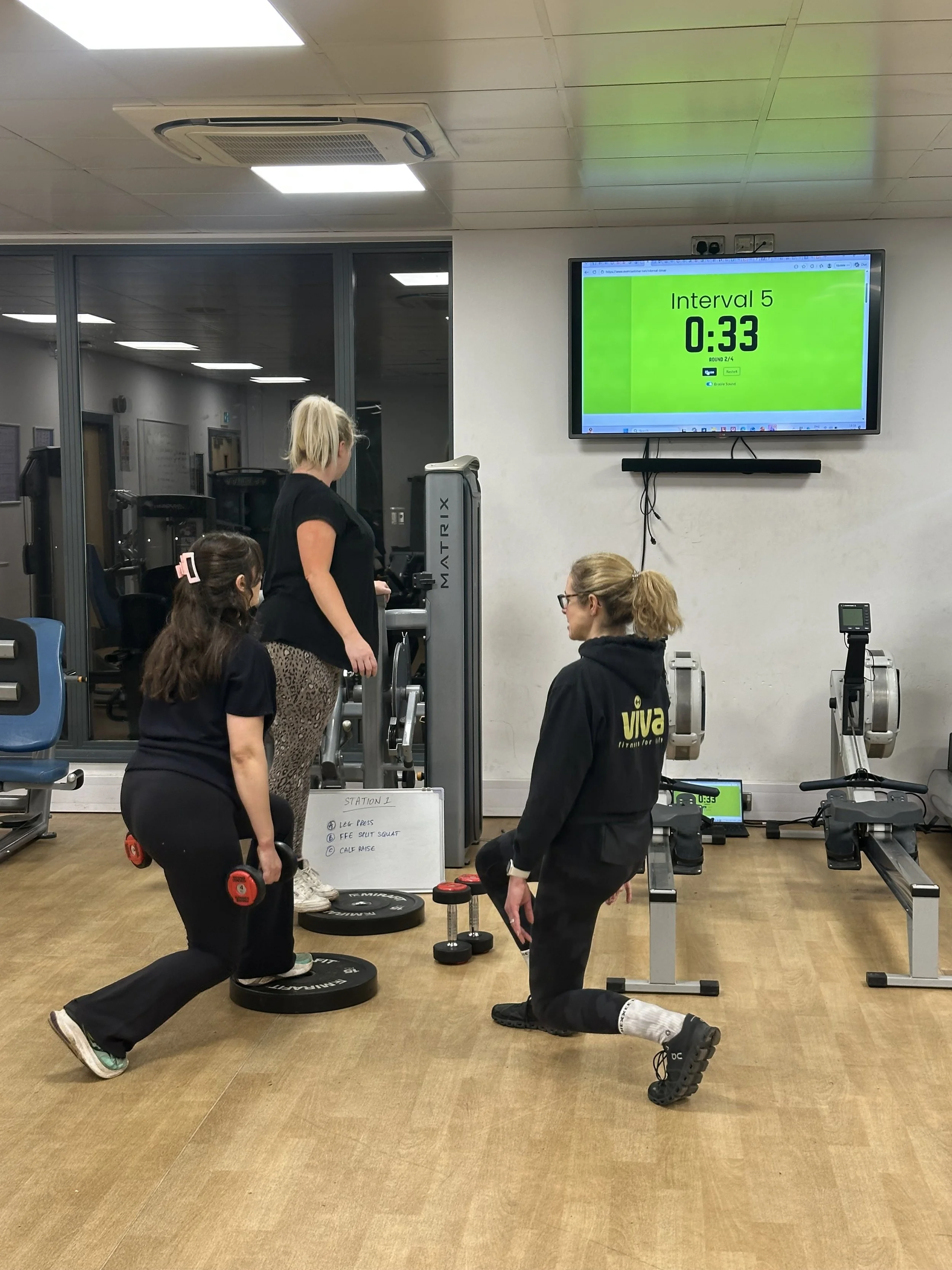 Two women, one kneeling and one standing, lifting dumbbells during a workout in a gym with muscular leg presses and a green digital timer on the wall. Other people are visible in the background.