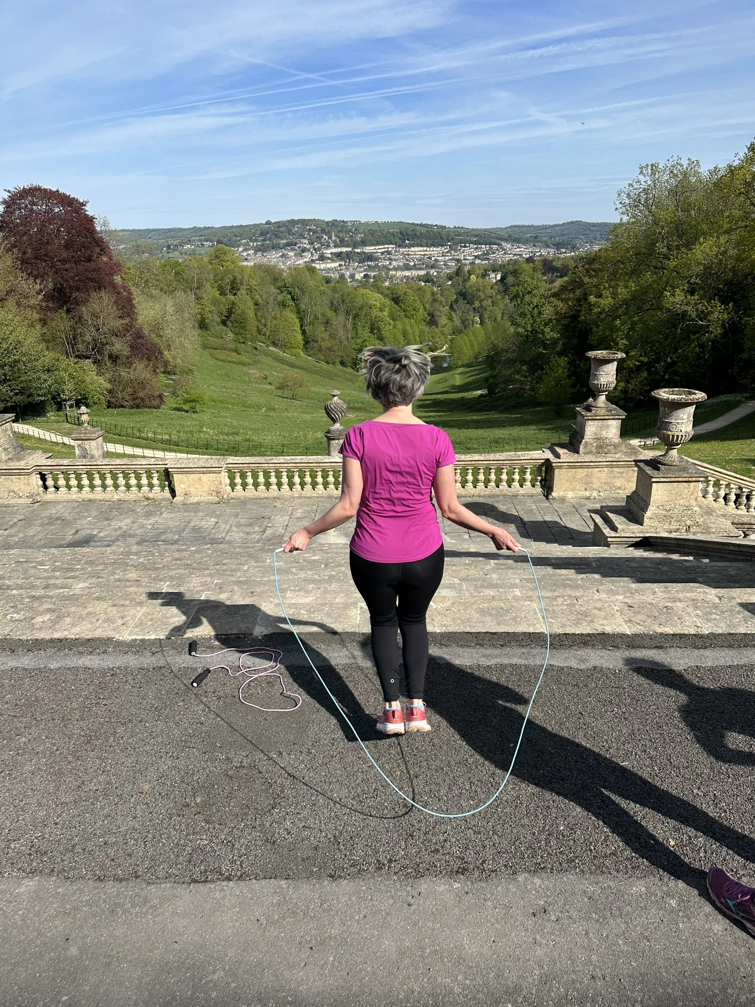 A woman in a pink top and black pants skipping rope on a stone-paved outdoor area overlooking a green valley with trees, a town, and hills on a clear day.