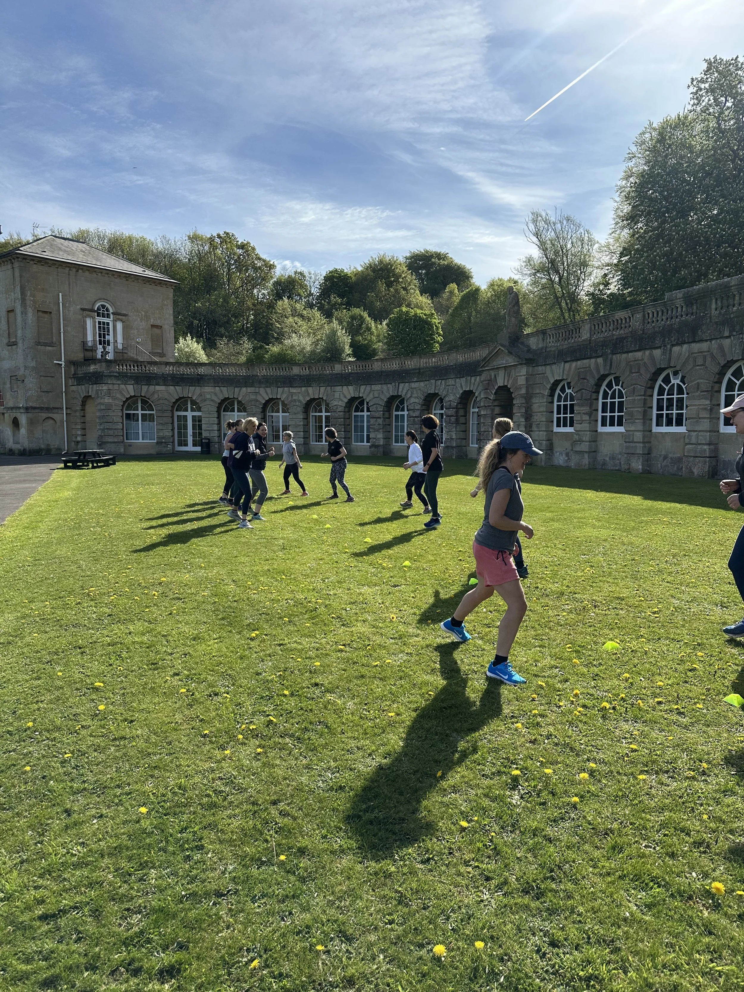 People running and walking on a grassy field in front of a stone building with arched windows, surrounded by trees under a bright blue sky.