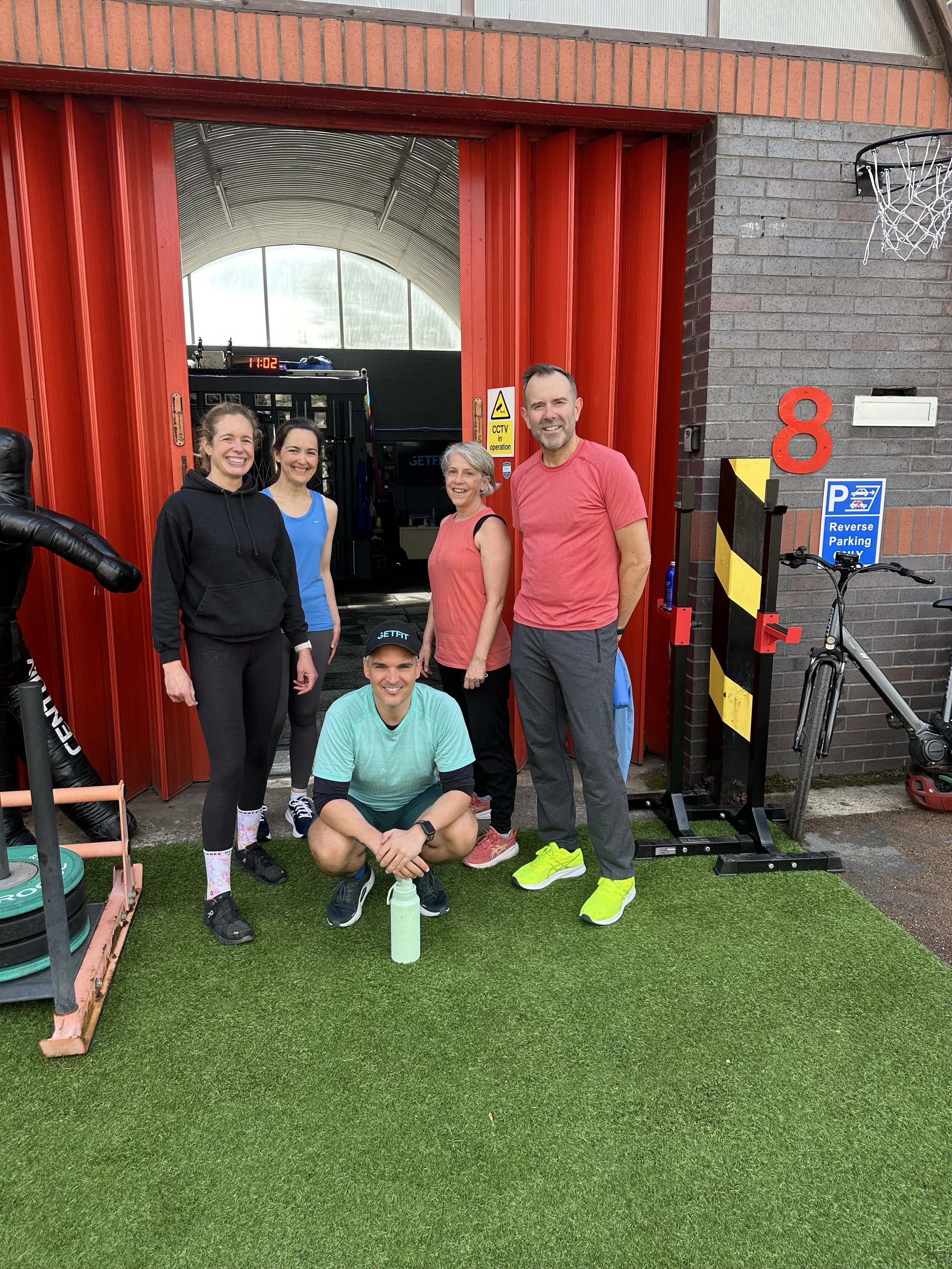 A group of five people, three women and two men, smiling and posing after a workout in a gym or fitness center, standing on artificial grass outdoors with gym equipment and a basketball hoop in the background.