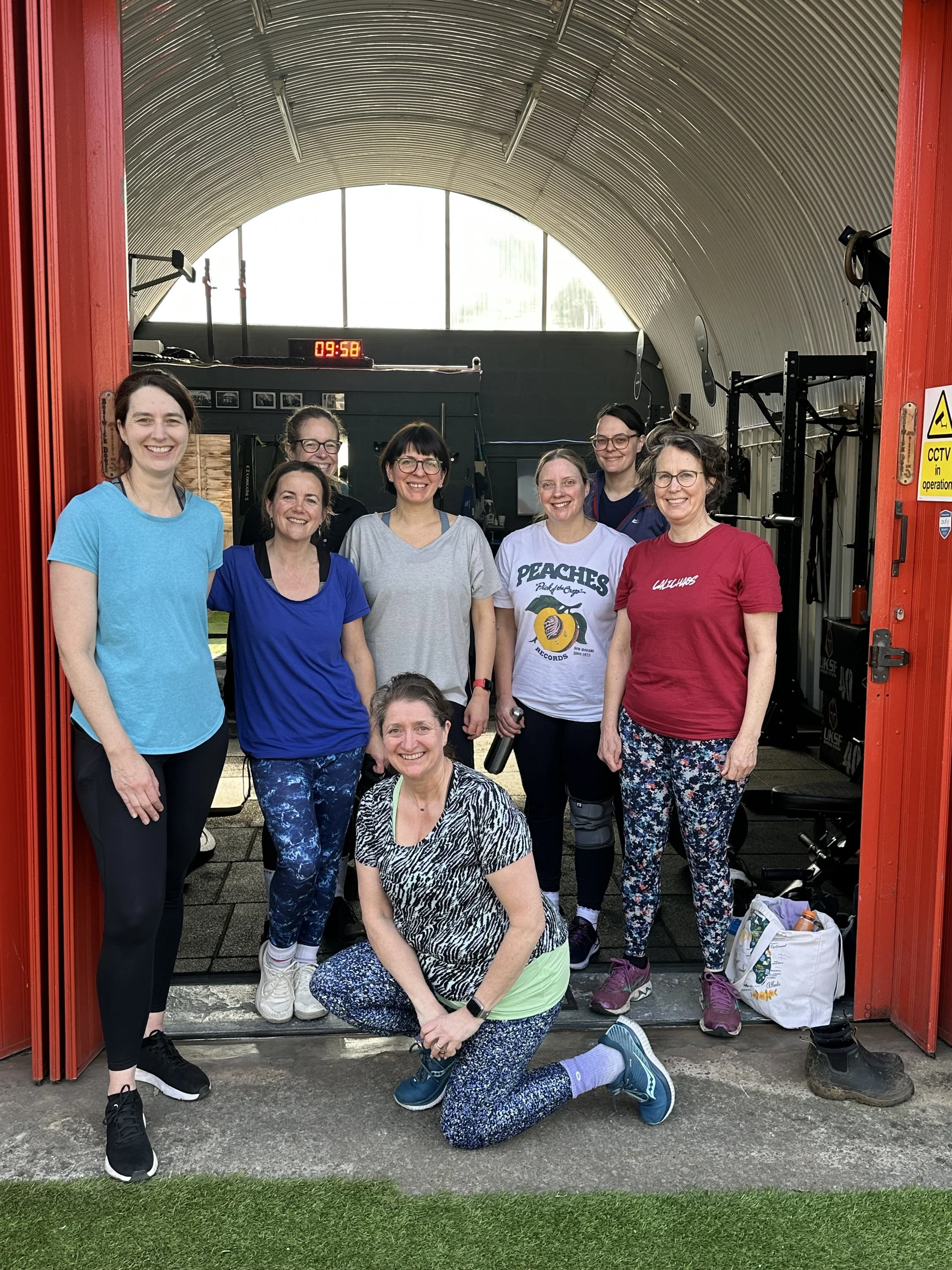 A group of nine women dressed in athletic clothing, posing together inside a red door frame of a small private gym in Bath.