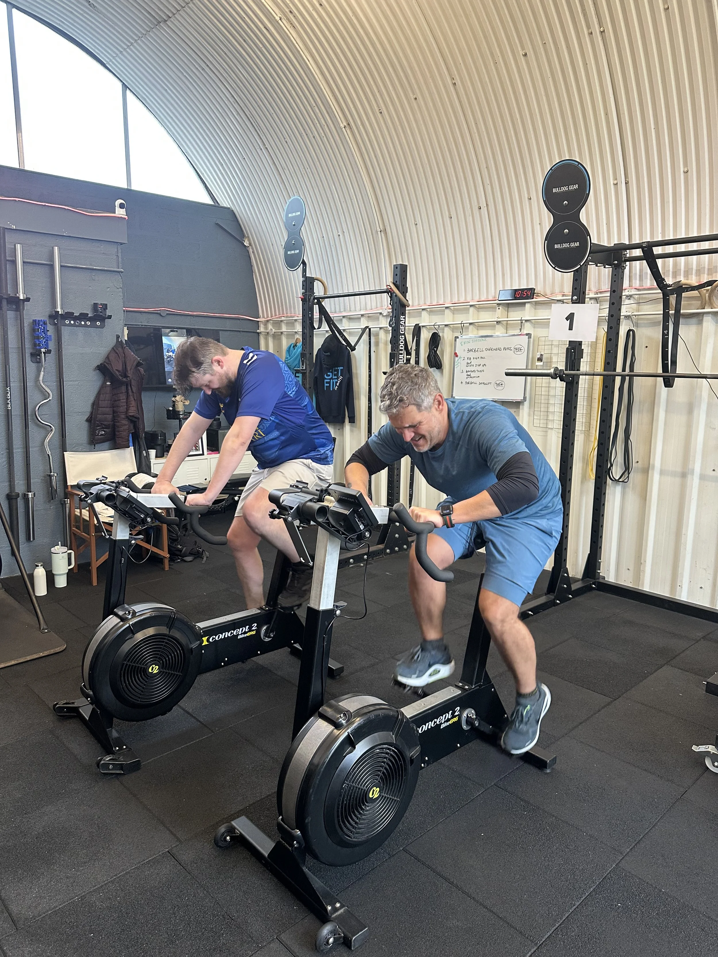 Two men working out on Concept2 indoor cycling machines in a gym, with one man wearing a blue shirt and the other wearing a gray shirt.