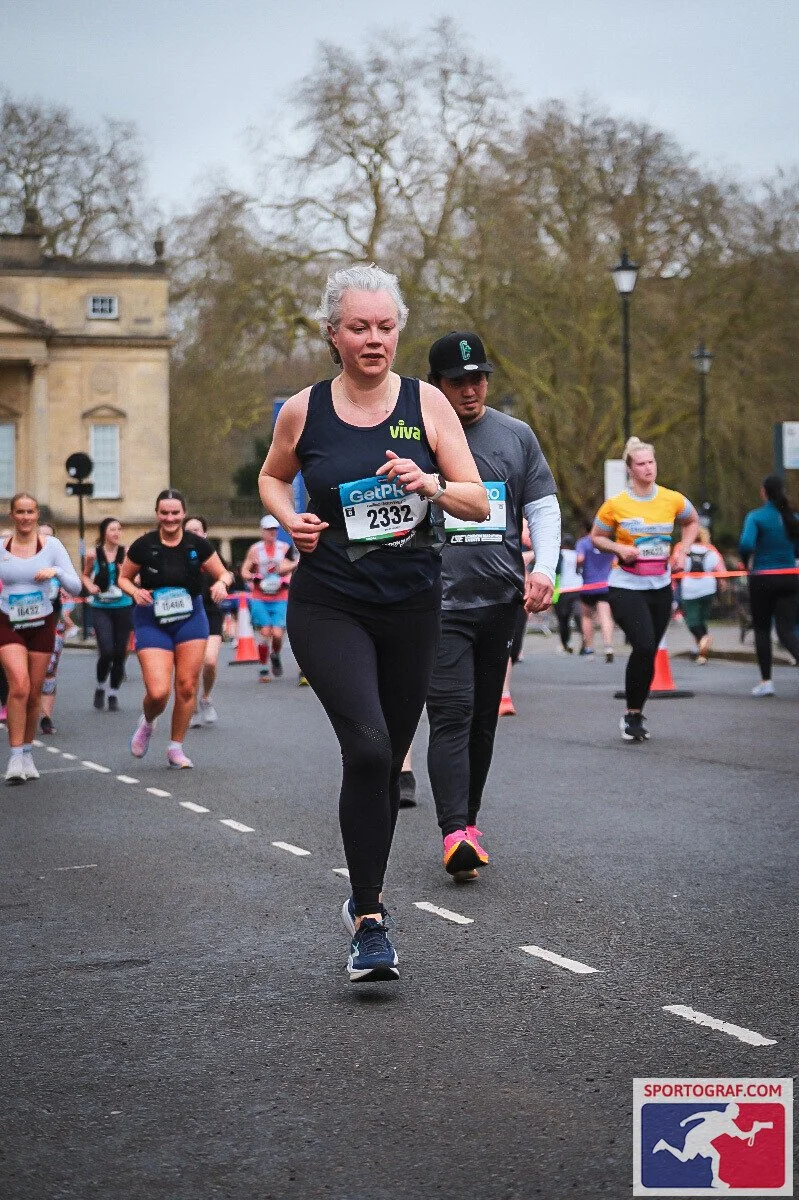 A group of runners participating in a race on a city street, with a woman in the foreground checking her watch, wearing a black tank top with 'Viva' on it, and a diverse group of runners behind her.