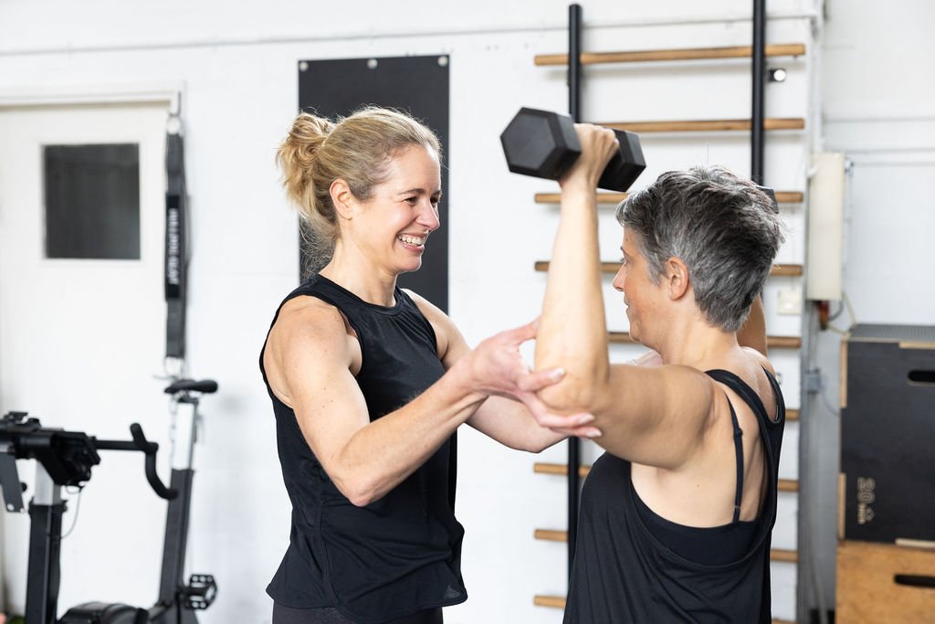 Two women in workout clothes at a gym, one helping the other with a dumbbell exercise, both smiling.