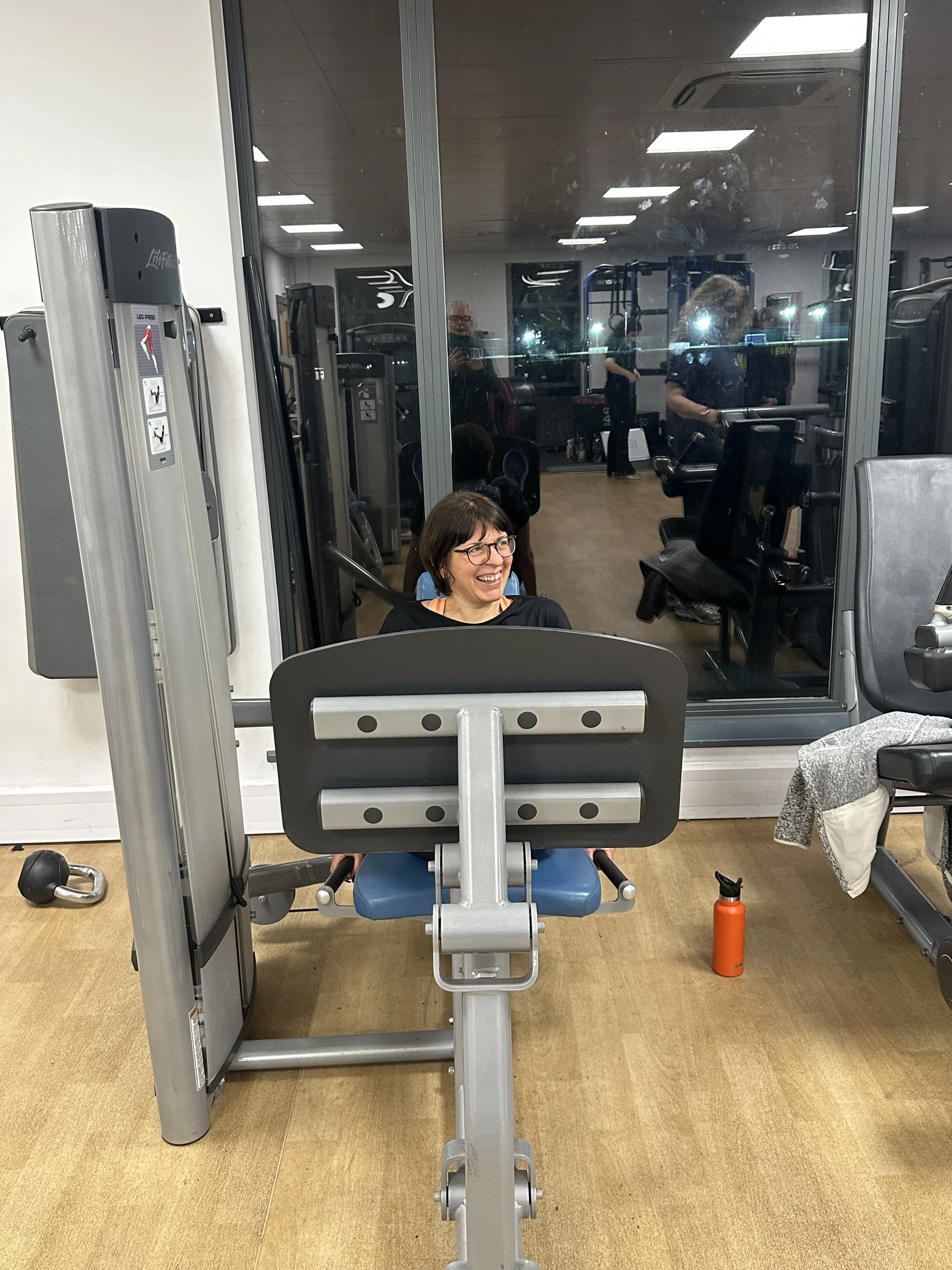 A woman in glasses and a black shirt sitting on exercise equipment in a gym, smiling. The gym has various machines and a large window with people visible through the glass.