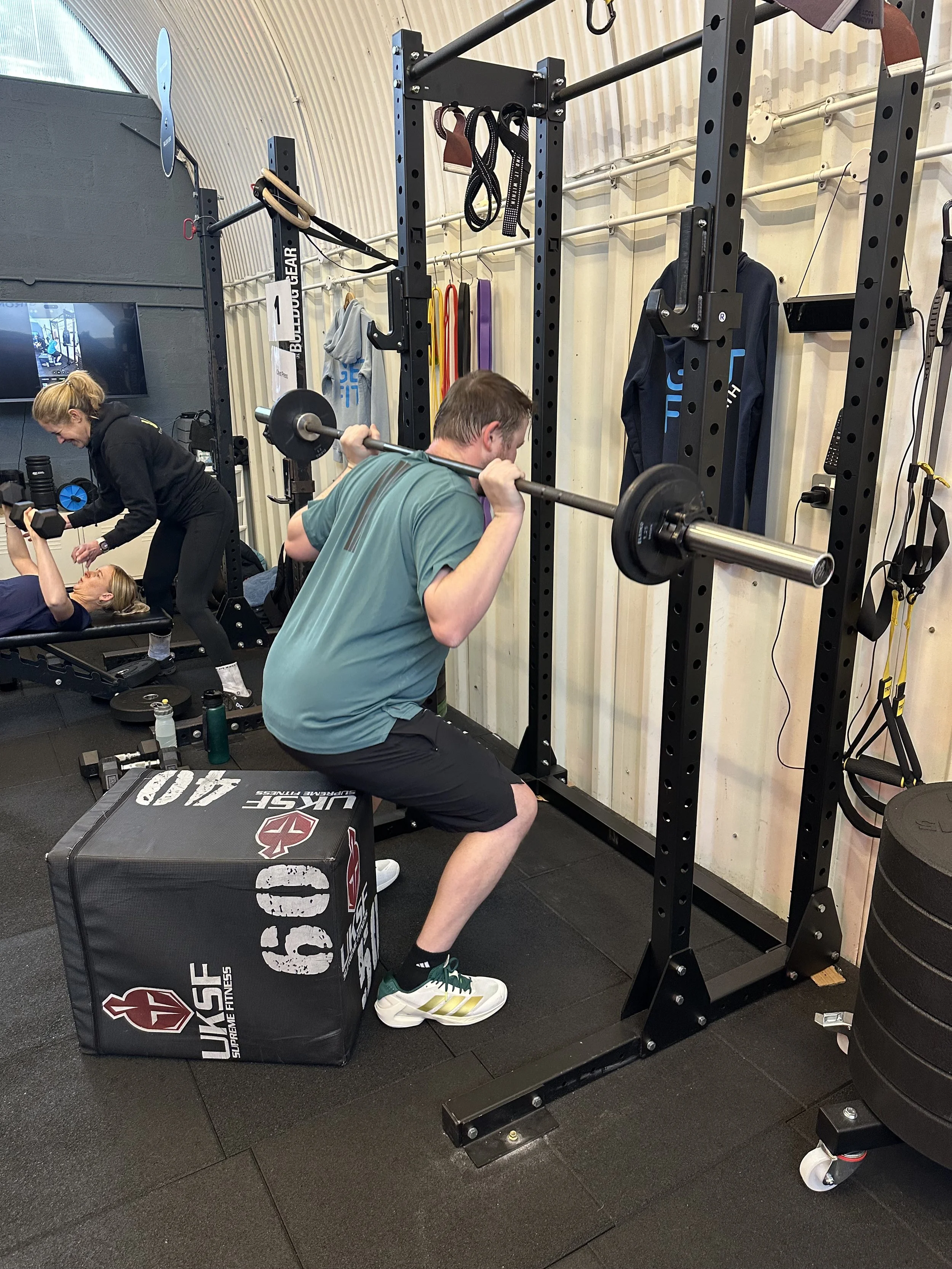 A man performing a barbell squat in a gym, with a woman assisting and a woman lying on a bench nearby.
