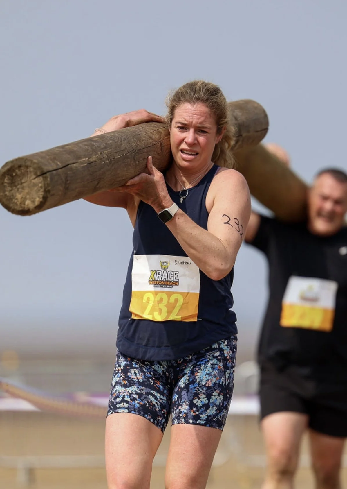 Woman participating in a race, carrying a large wooden log on her shoulder, wearing a navy blue tank top with race bib number 232 and patterned shorts, with a man in the background also carrying a log.