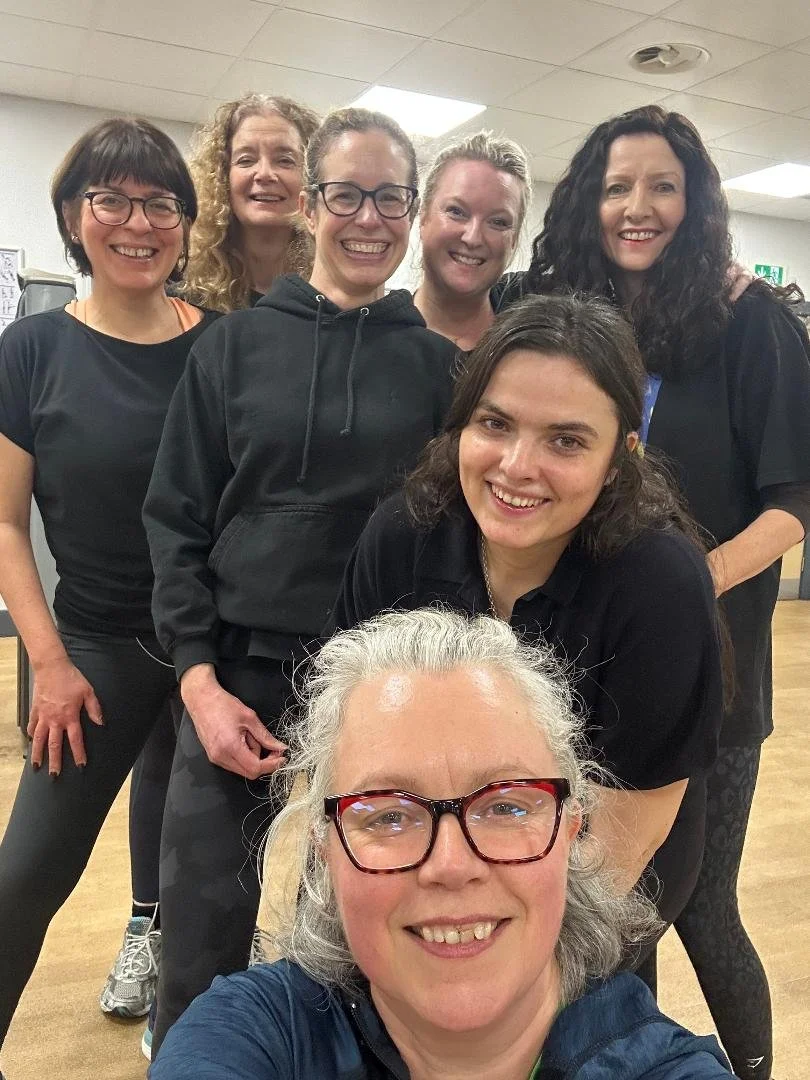 Six women in casual attire smiling and posing for a group selfie in a small private gym