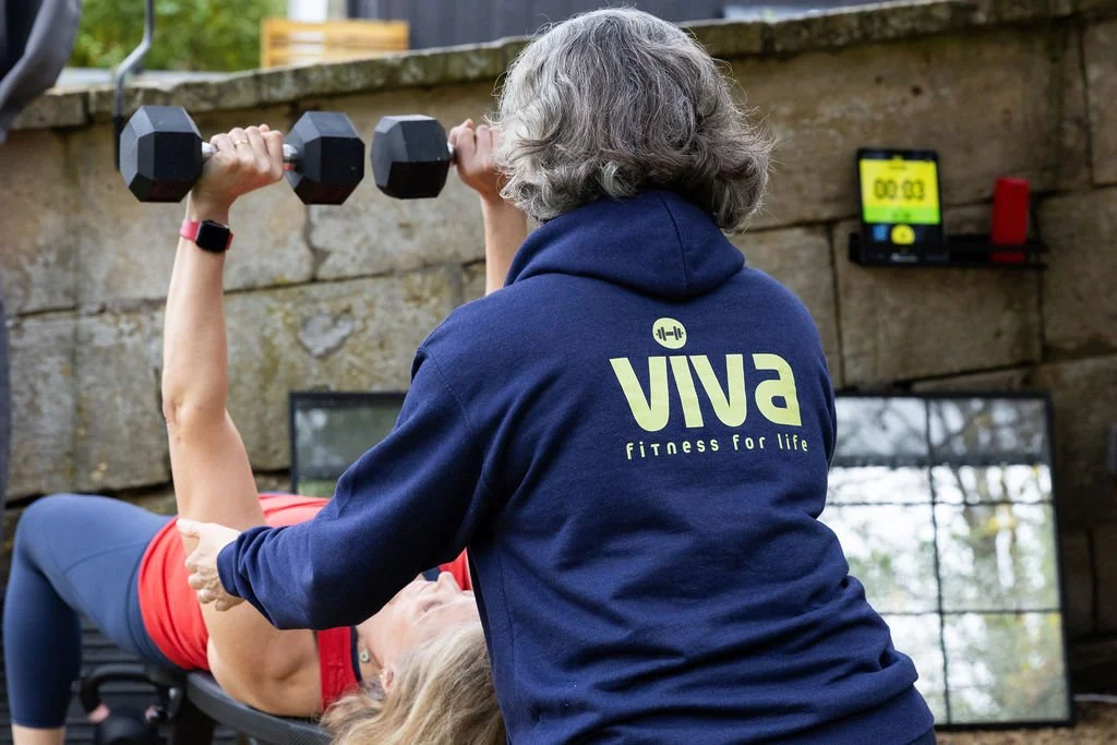 A woman in a blue sweatshirt with the VIVA fitness logo assists a woman exercising with dumbbells outdoors, with a stopwatch and mirror in the background.