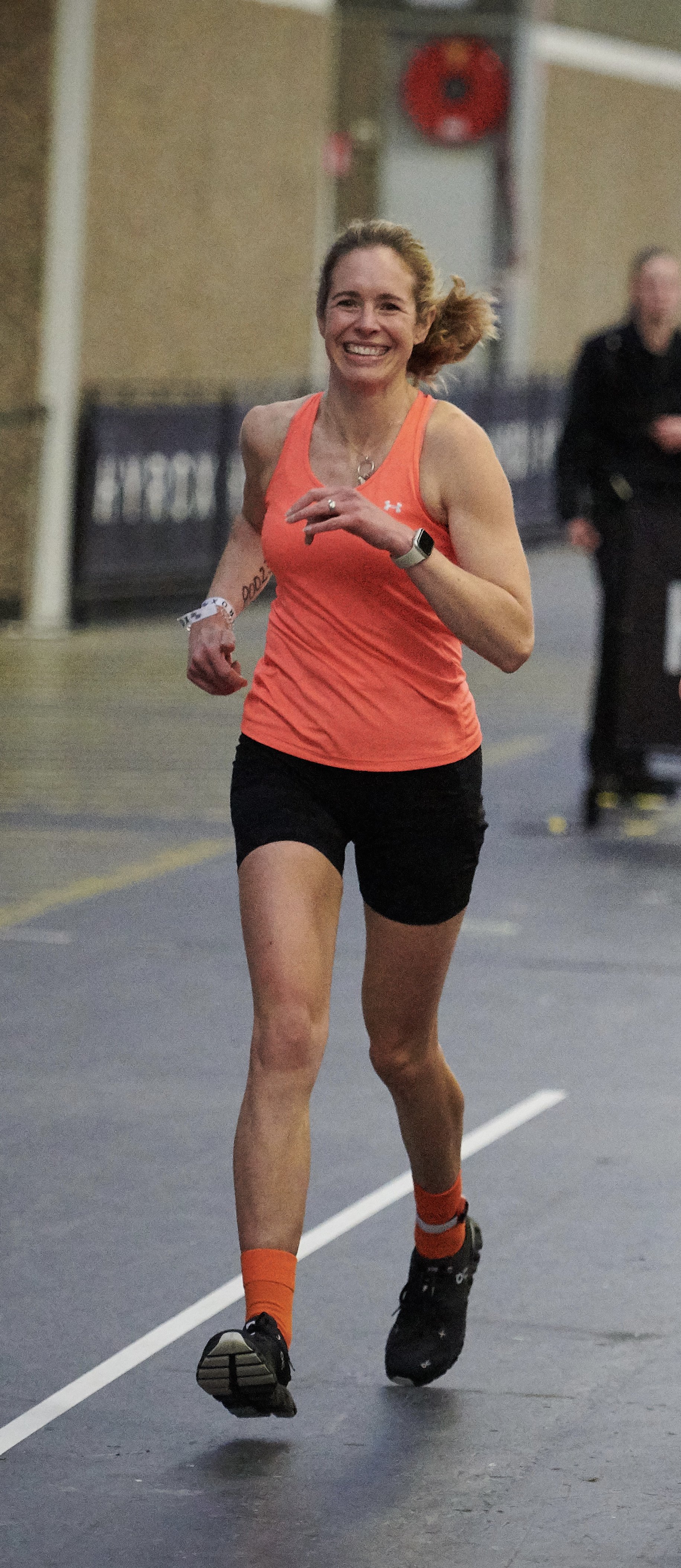 A woman running indoors, wearing a bright orange tank top, black shorts, orange socks, and black running shoes, smiling as she runs.