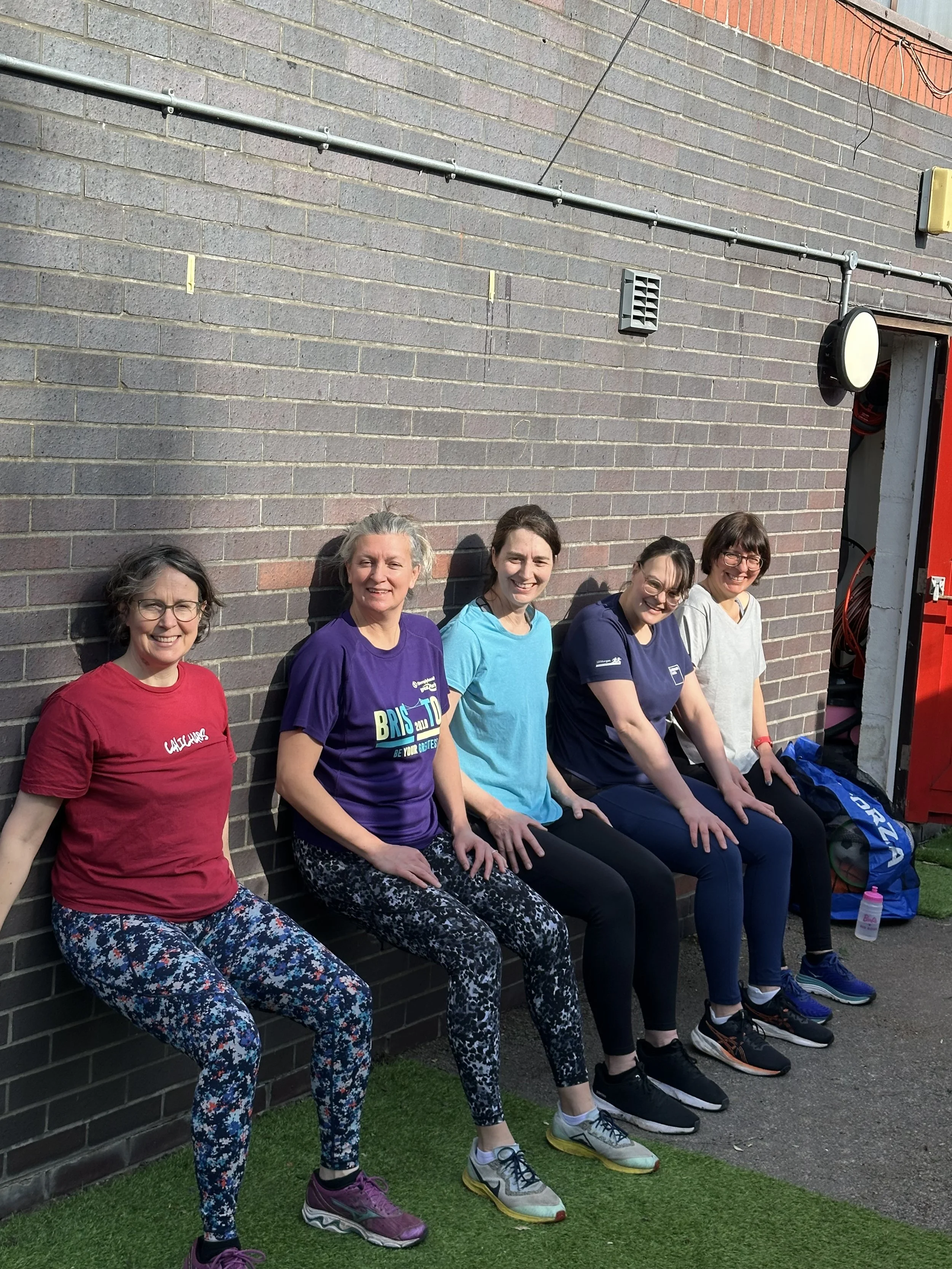 A group of five women sitting on a bench against a brick wall during a workout, smiling. They are wearing colorful workout clothes and sneakers.