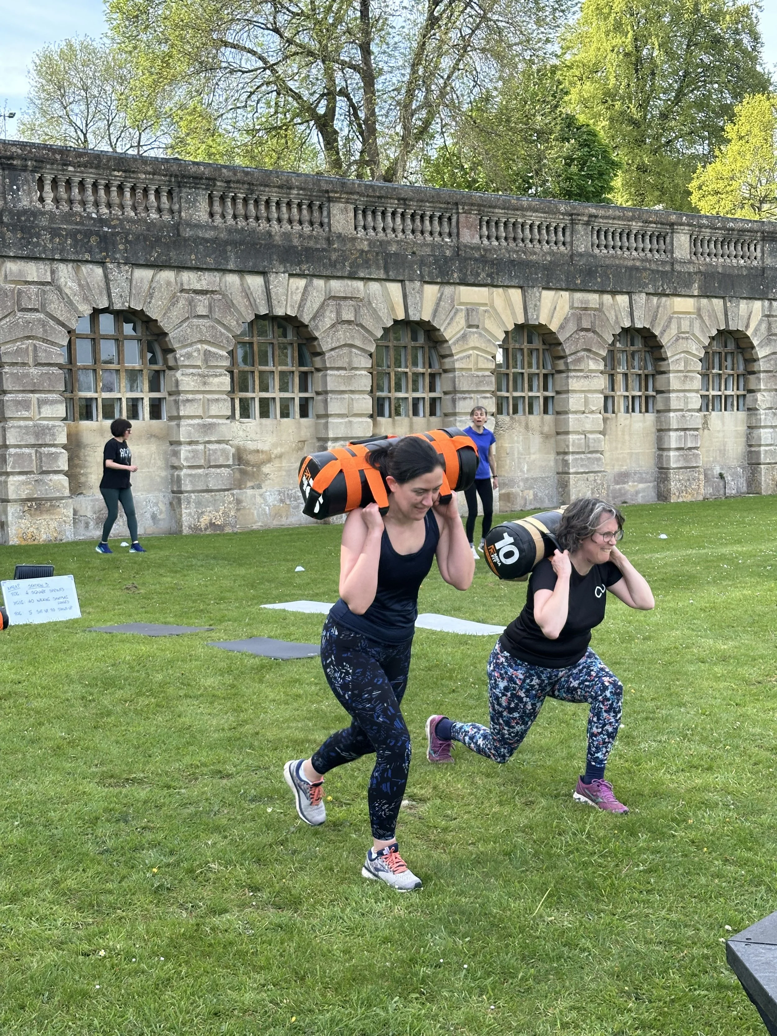 Two women are running outdoors on a grassy area carrying heavy bags on their shoulders during a fitness event, with a stone building and trees in the background.