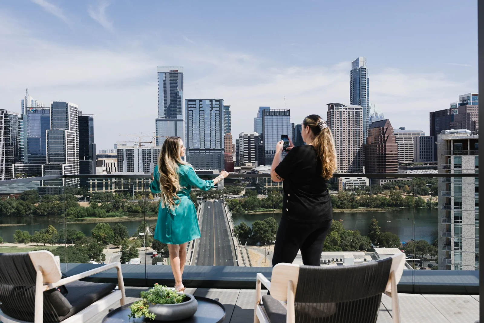 Two women are on a balcony with a view of a city skyline featuring tall skyscrapers. One woman in a turquoise dress is posing, while the other in black is taking a photo.