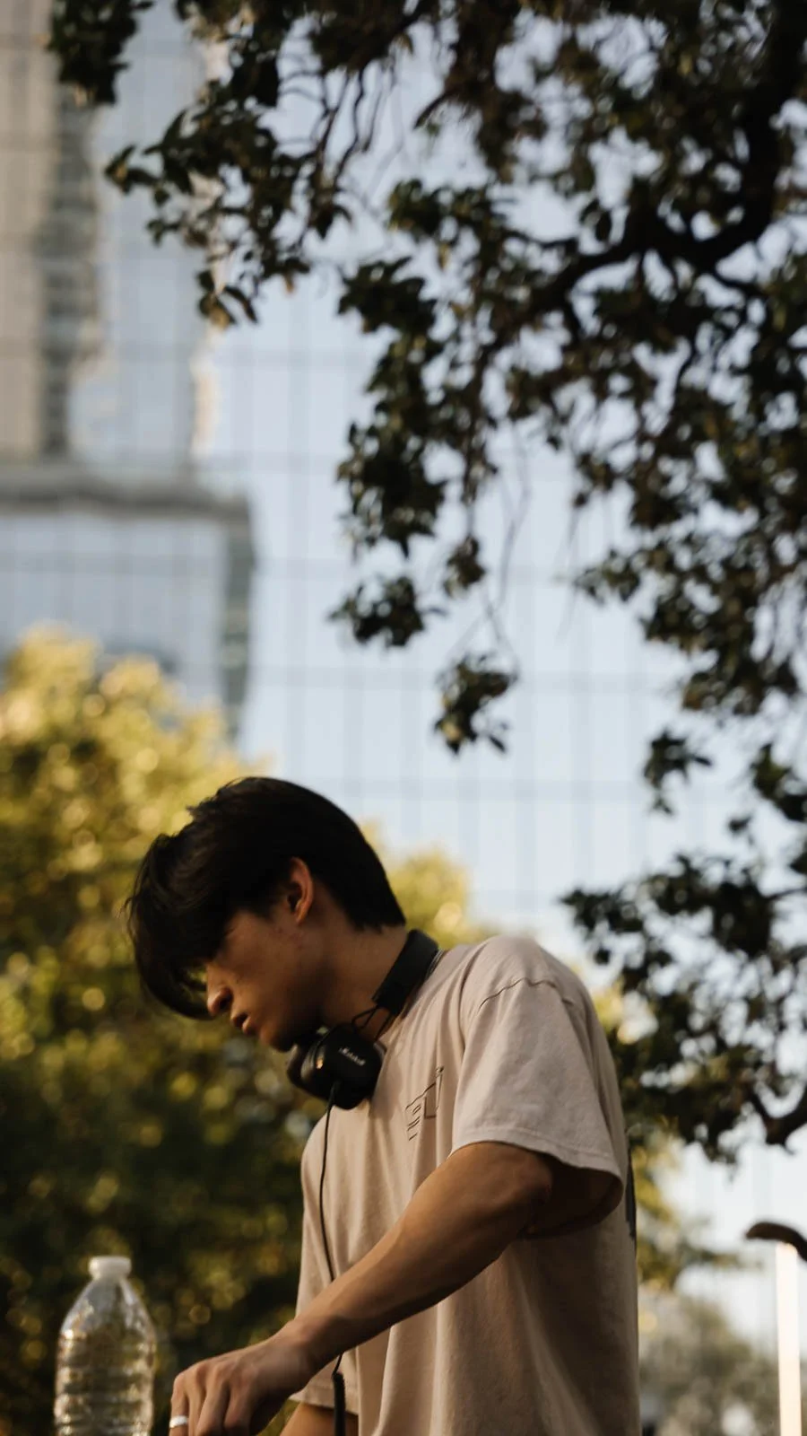 Young man with black headphones around neck, wearing beige t-shirt, standing outdoors with trees and urban buildings in the background, looking down.