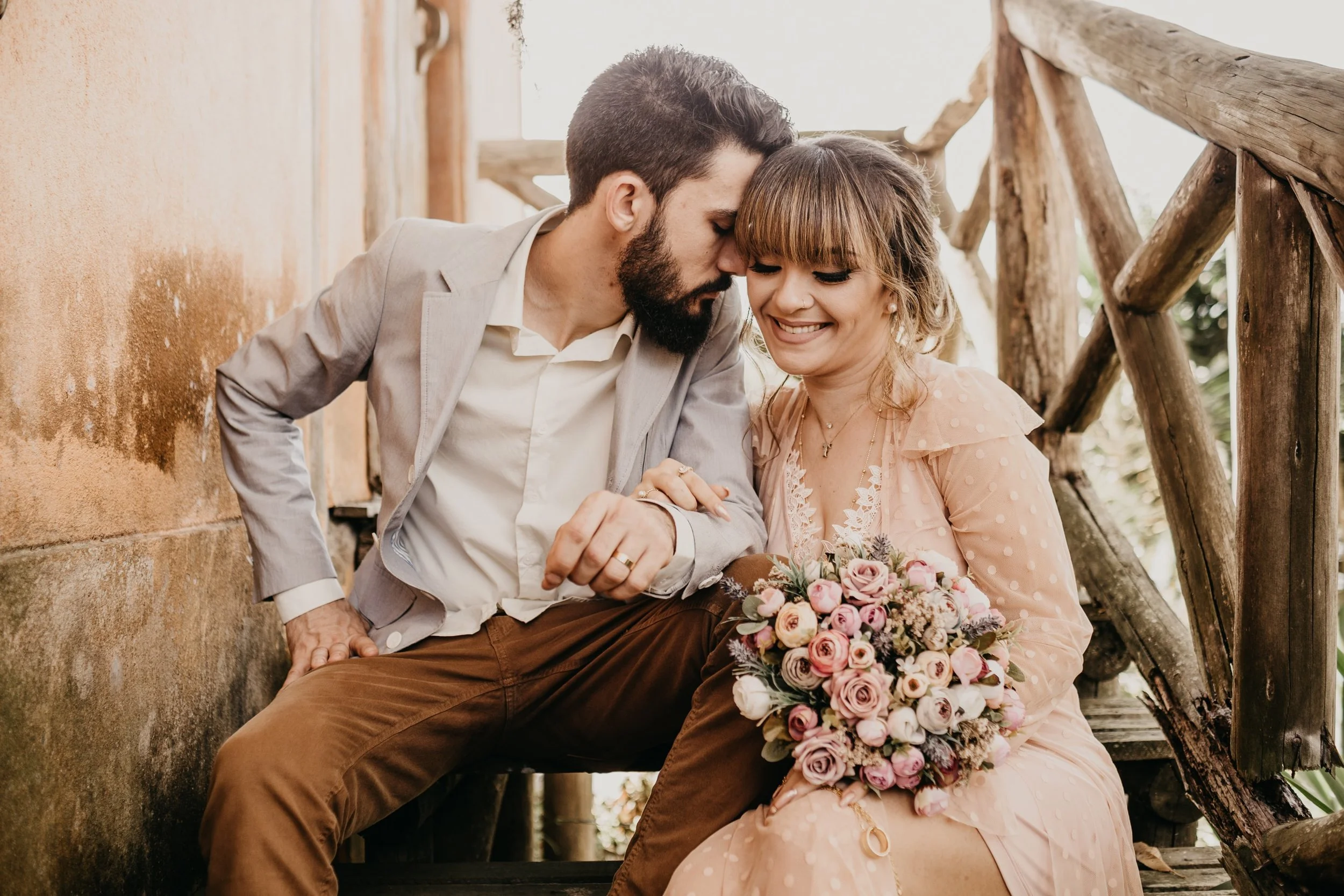 A man and woman sitting closely together on a wooden staircase with a rustic railing, sharing a tender moment. The woman holds a bouquet of pink and white roses, and they are both smiling with their eyes closed, touching foreheads.
