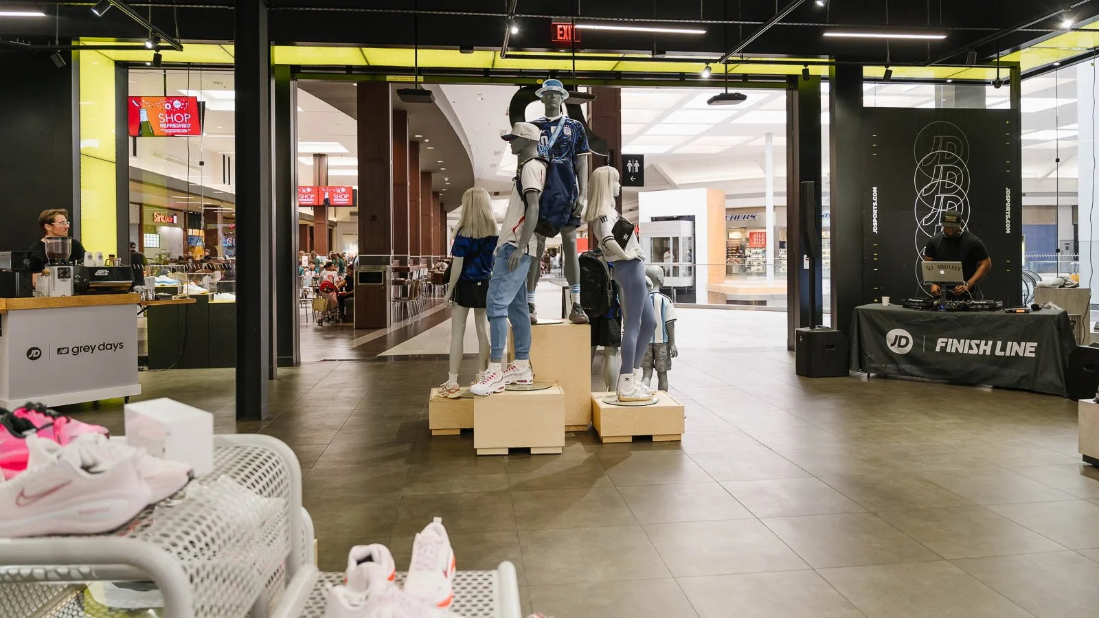 Inside a shopping mall with a display of mannequins dressed in athletic wear and sneakers at a Nike store, a DJ set up with a DJ on the right side, and a kiosk on the left side.