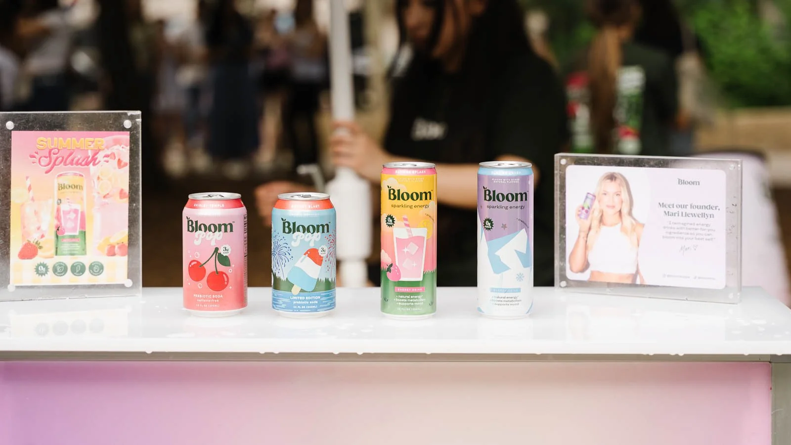 Display of Bloom beverages including cans of prebiotic soda and sparkling energy drinks on a white table at an outdoor event, with informational signs about the founder Mari Llewellyn and the products.