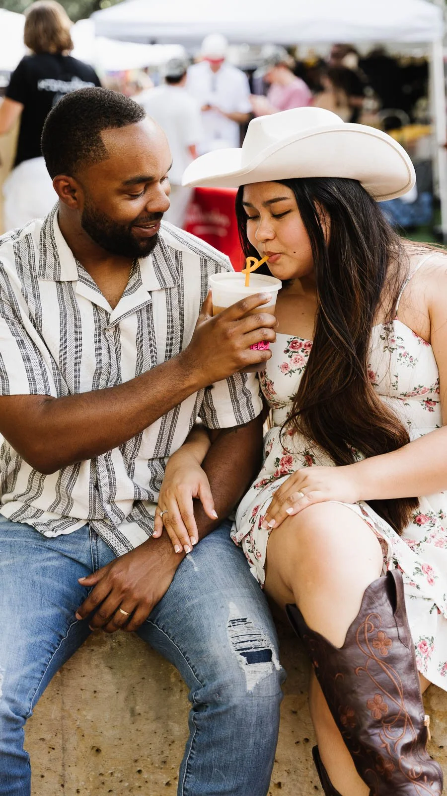 A man and woman sharing a drink at an outdoor festival or fair, with food stalls and people in the background.